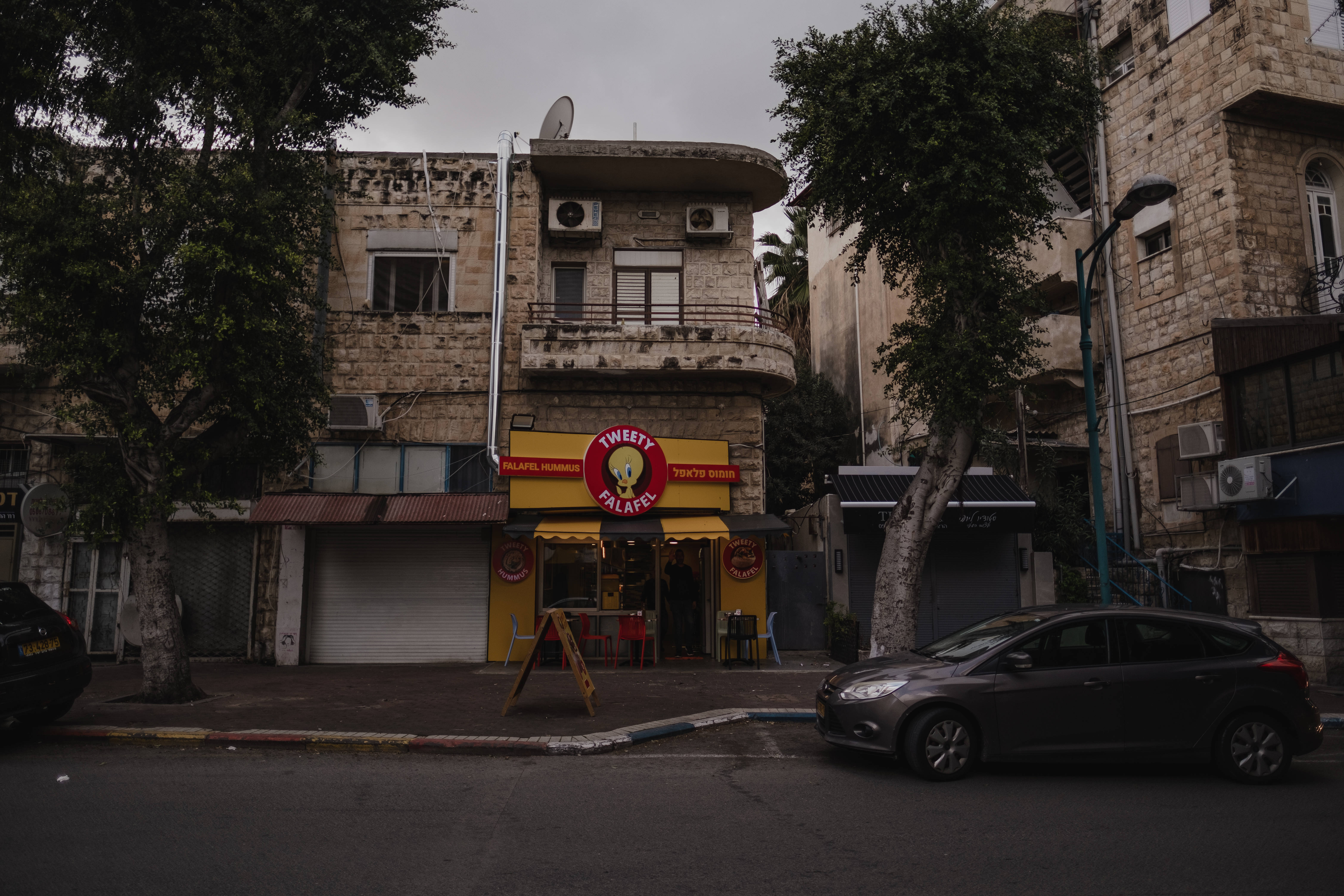 Israel 桌布 壁紙 — 一條狹窄的街道兩旁是風化的石頭建築，在陰天之下有一間亮黃色的沙威瑪店。