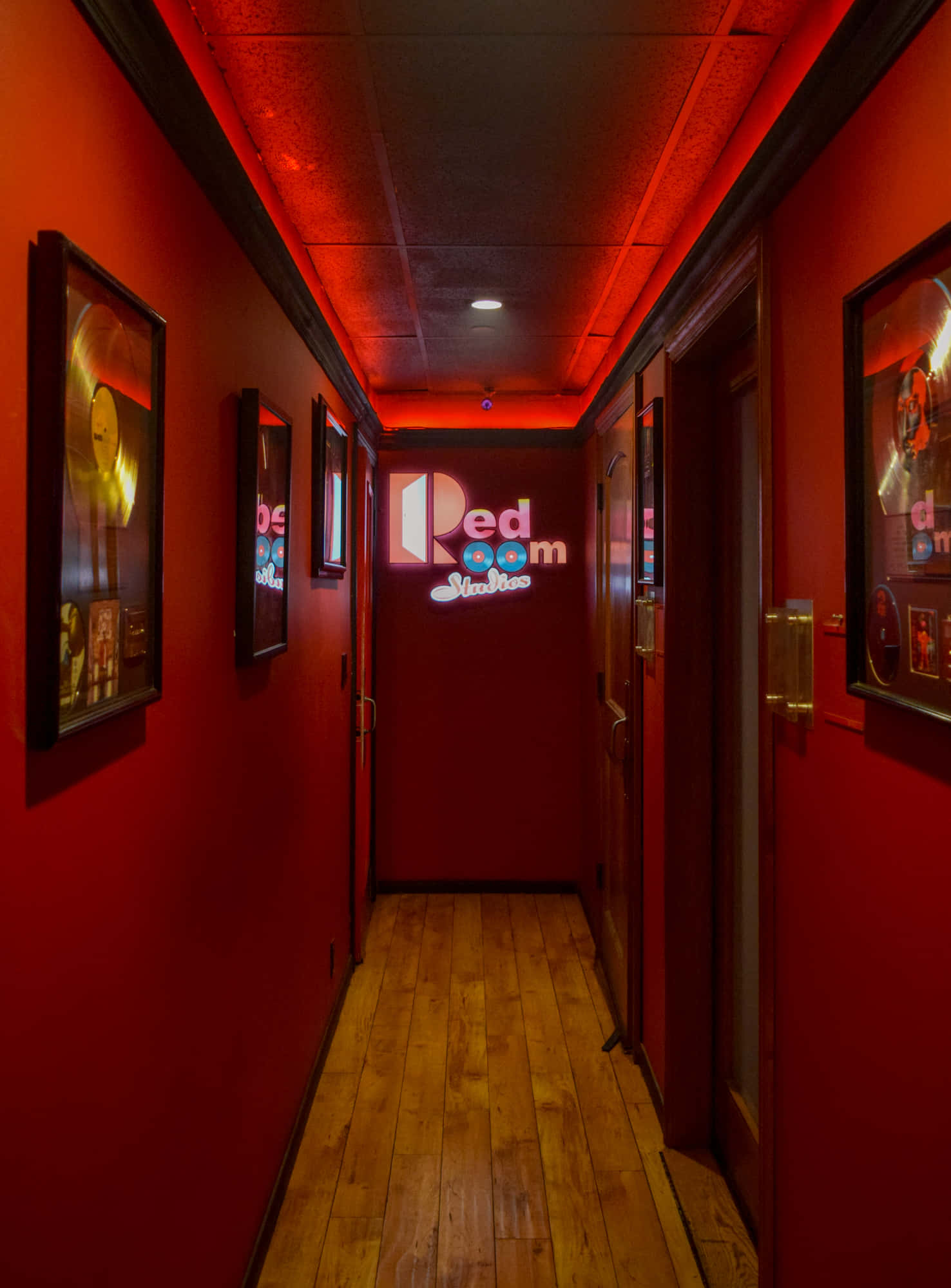 Red Room 桌布 壁紙 — 一條狹窄的走廊沐浴在深紅色的燈光中，木地板和裱框海報通向一個發光的霓虹燈招牌，上面寫著 Red Room Studios。