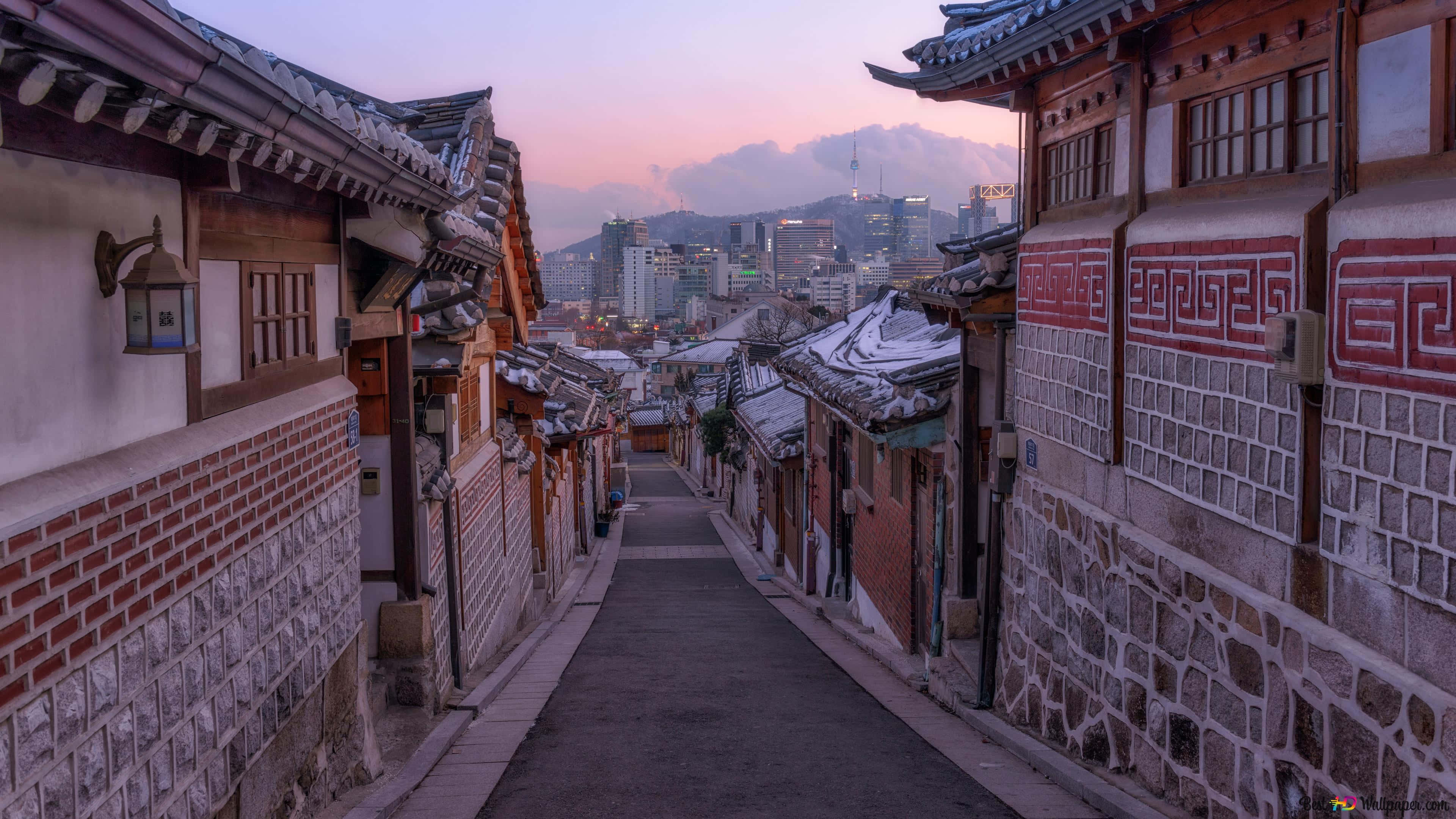 南韓 桌布 壁紙 — 一條狹窄的雪巷，兩旁是傳統韓國韓屋，暮色中遠方可見現代城市天際線。