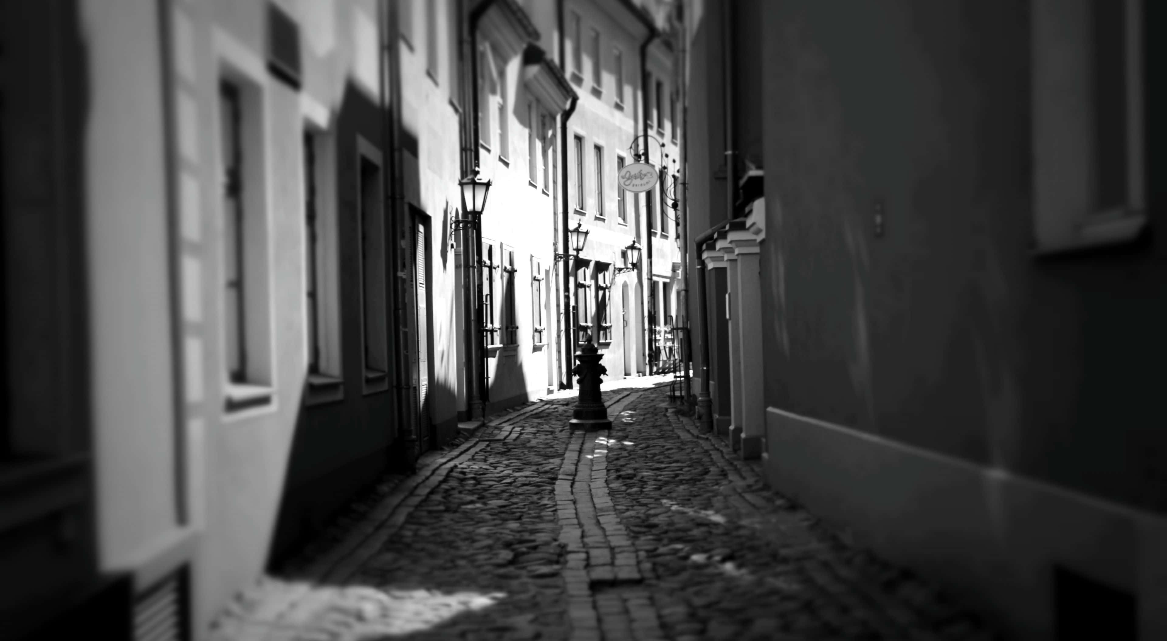 Black And White Street 桌布 壁紙 — 一條狹窄的鵝卵石小巷，位於古老的歐洲建築之間，沐浴在戲劇性的黑白光線中。