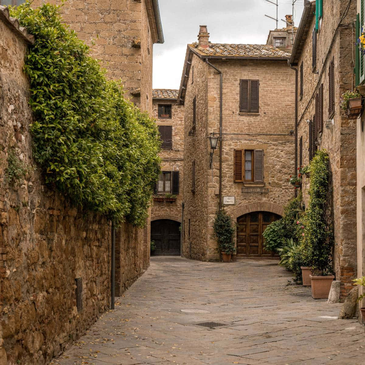 Tuscany 桌布 壁紙 — 一條狹窄的鵝卵石小巷，位於古老的歐洲村莊，兩旁是風化的石頭建築和茂密的綠色植物。