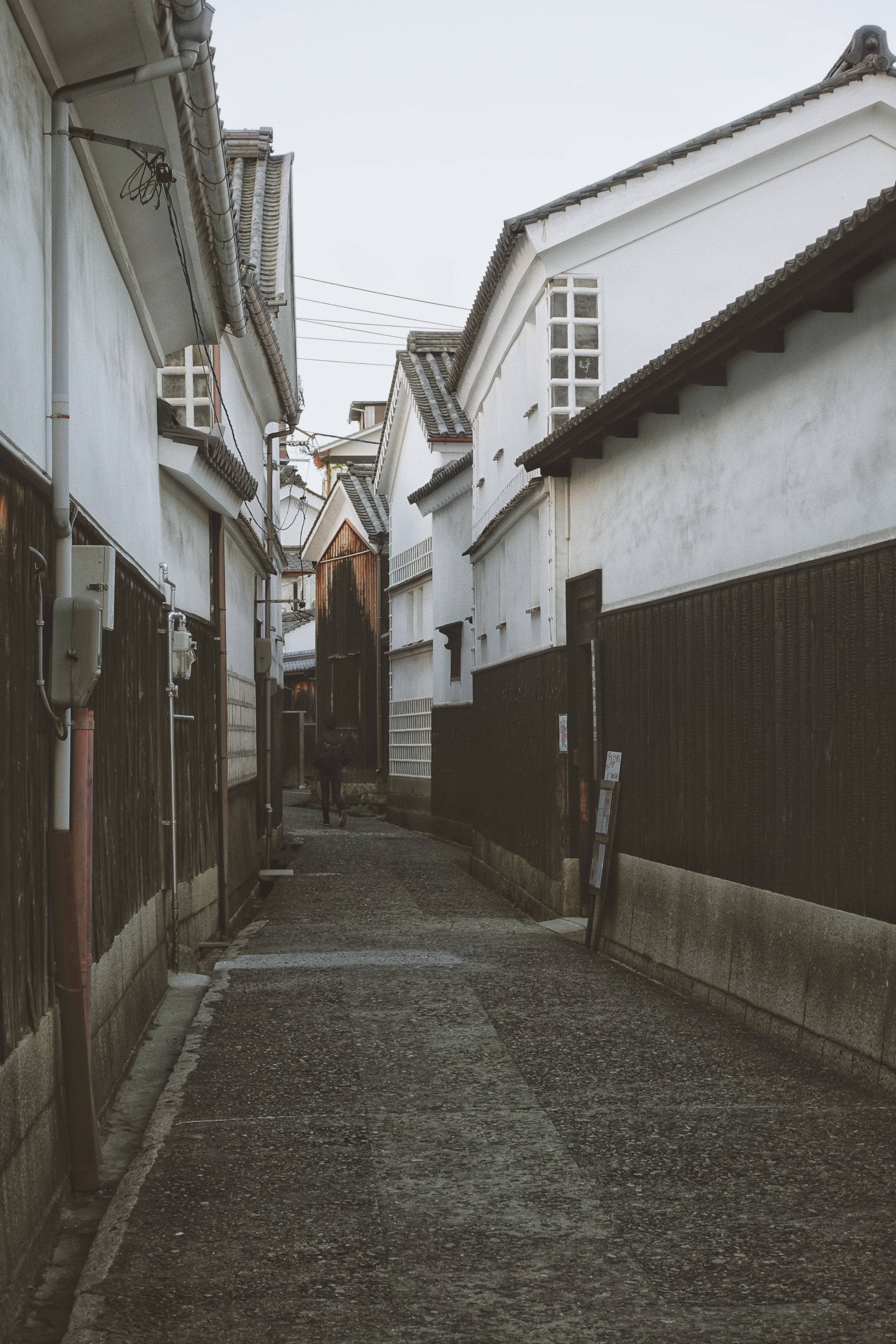 Street 桌布 壁紙 — 一條狹窄的鵝卵石小巷，兩旁是傳統的白牆日本房屋和深色木柵欄，天空多雲。