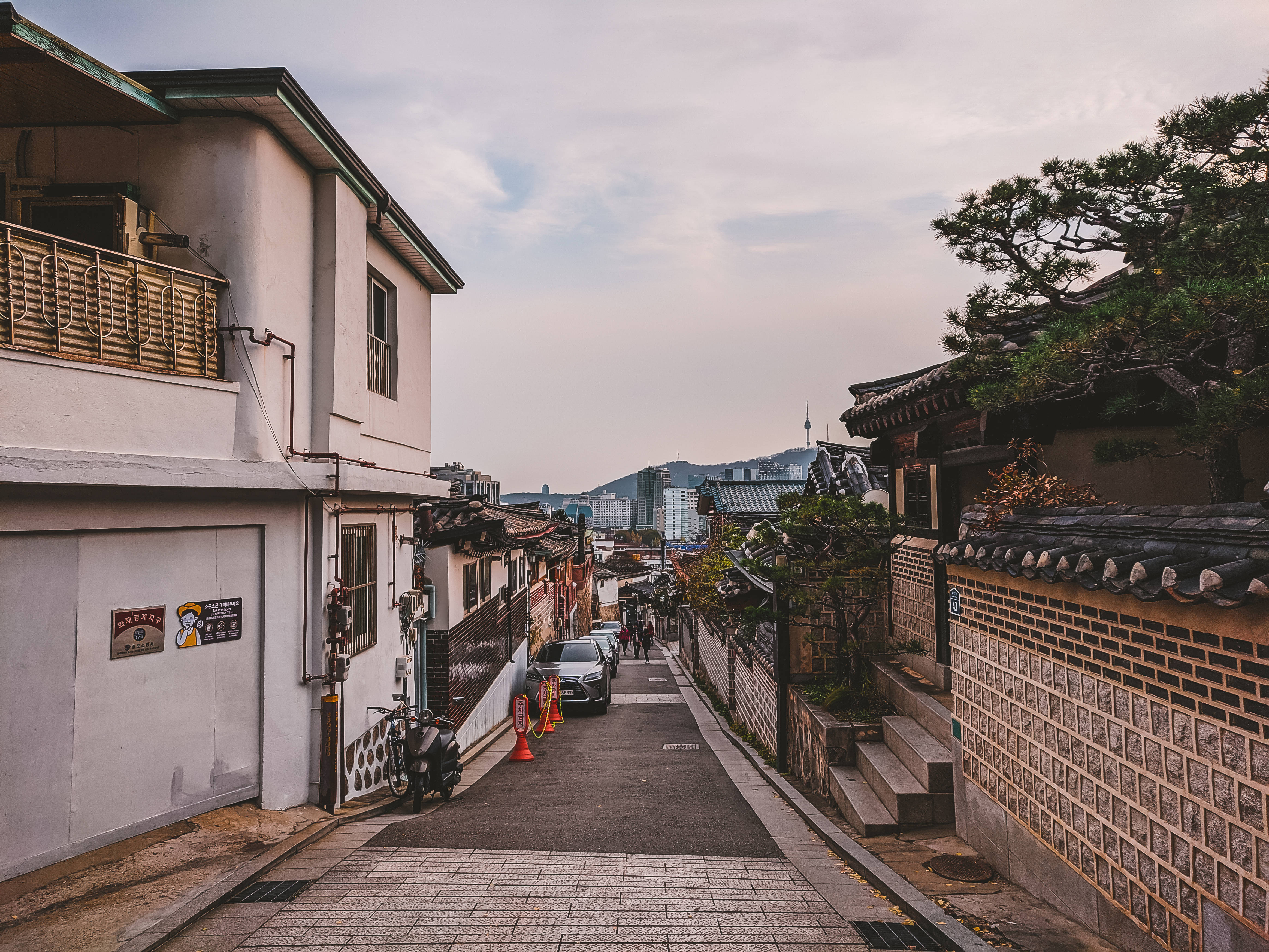 Seoul 桌布 壁紙 — 一條狹窄的鵝卵石小巷在傳統韓國社區中，在柔和暮色天空下，在瓦頂和石牆之間向上傾斜。