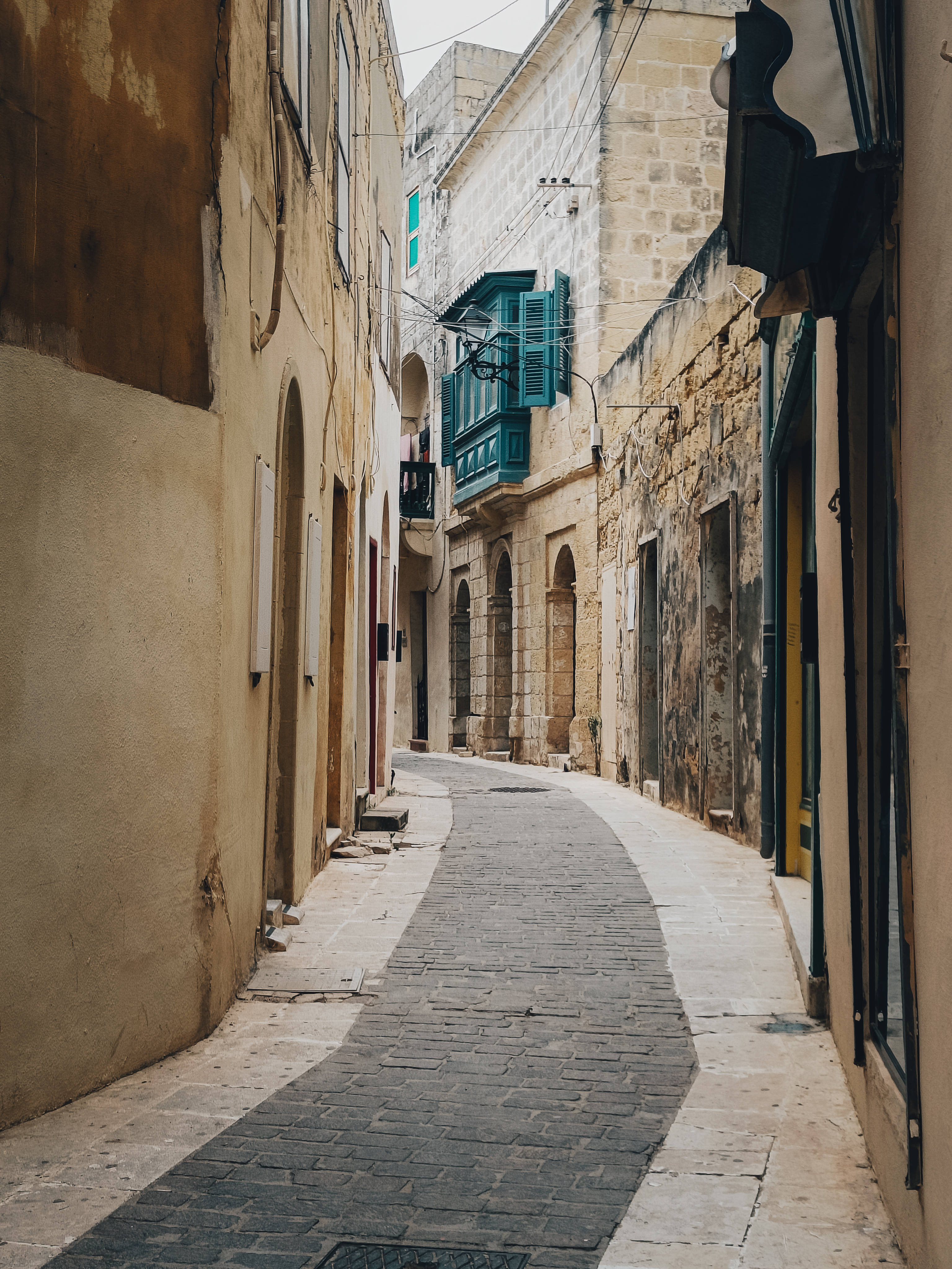Malta 桌布 壁紙 — 一條狹窄的鵝卵石小巷蜿蜒在風化的石建築之間，一個充滿活力的青綠色窗台花箱增添了一抹色彩。