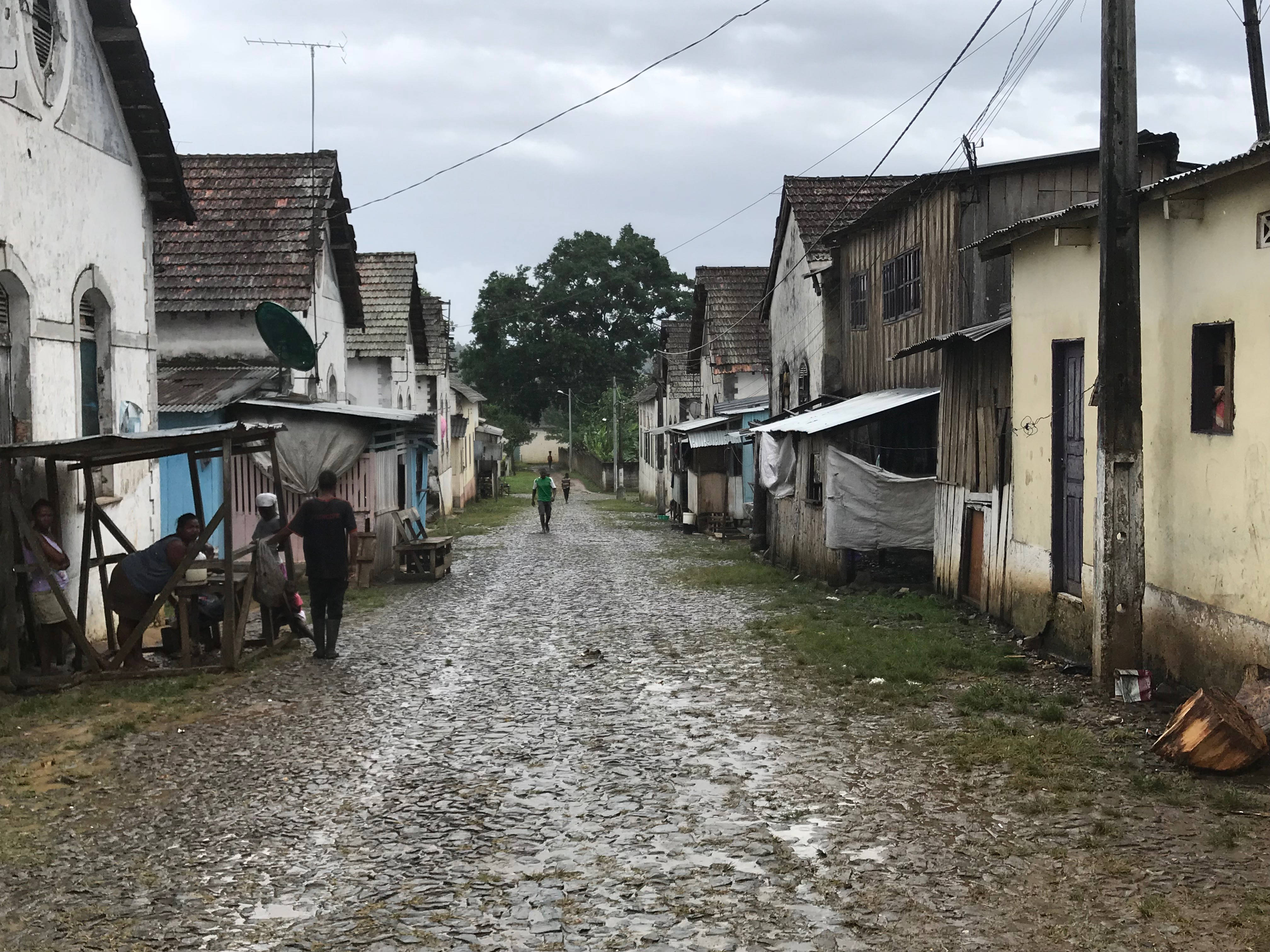 Road 桌布 壁紙 — 一條狹窄的鵝卵石街道，位於一個歷經風霜的社區，人們在多雲的日子裡在臨時搭建的遮陽篷下行走和坐著。