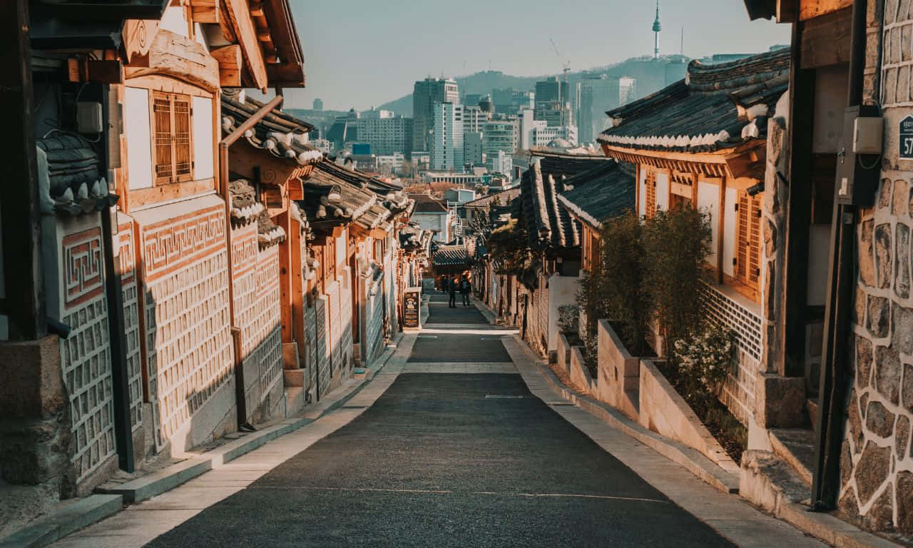 1280x768 桌布 壁紙 — 一條狹窄的鵝卵石街道，兩旁是傳統的韓國韓屋，通向晴朗藍天下現代化的城市天際線。
