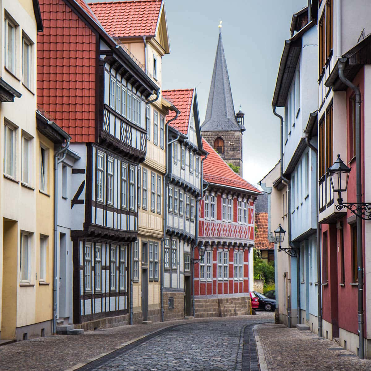 German 桌布 壁紙 — 一條狹窄的鵝卵石街道，兩旁是色彩繽紛的半木造房屋，通向陰天下的教堂尖塔。