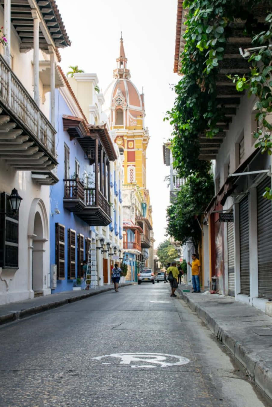 Colombia 桌布 壁紙 — 一條狹窄的鵝卵石街道，兩旁是色彩繽紛的殖民時期建築和茂密的綠植，通向一座充滿活力的教堂塔樓。