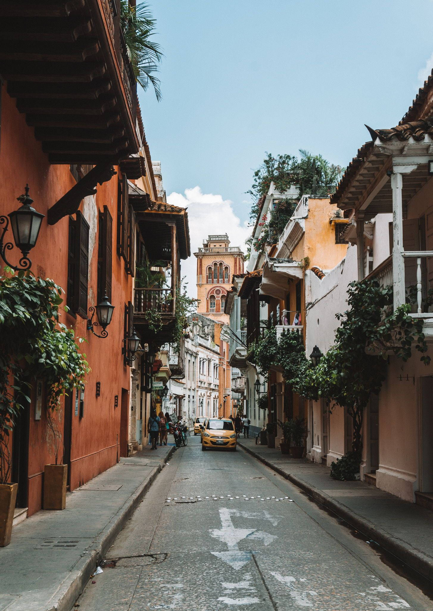 Colombia 桌布 壁紙 — 一條狹窄的鵝卵石街道，兩旁是色彩繽紛的殖民時期建築和茂密的綠植，上方是明亮的藍天。