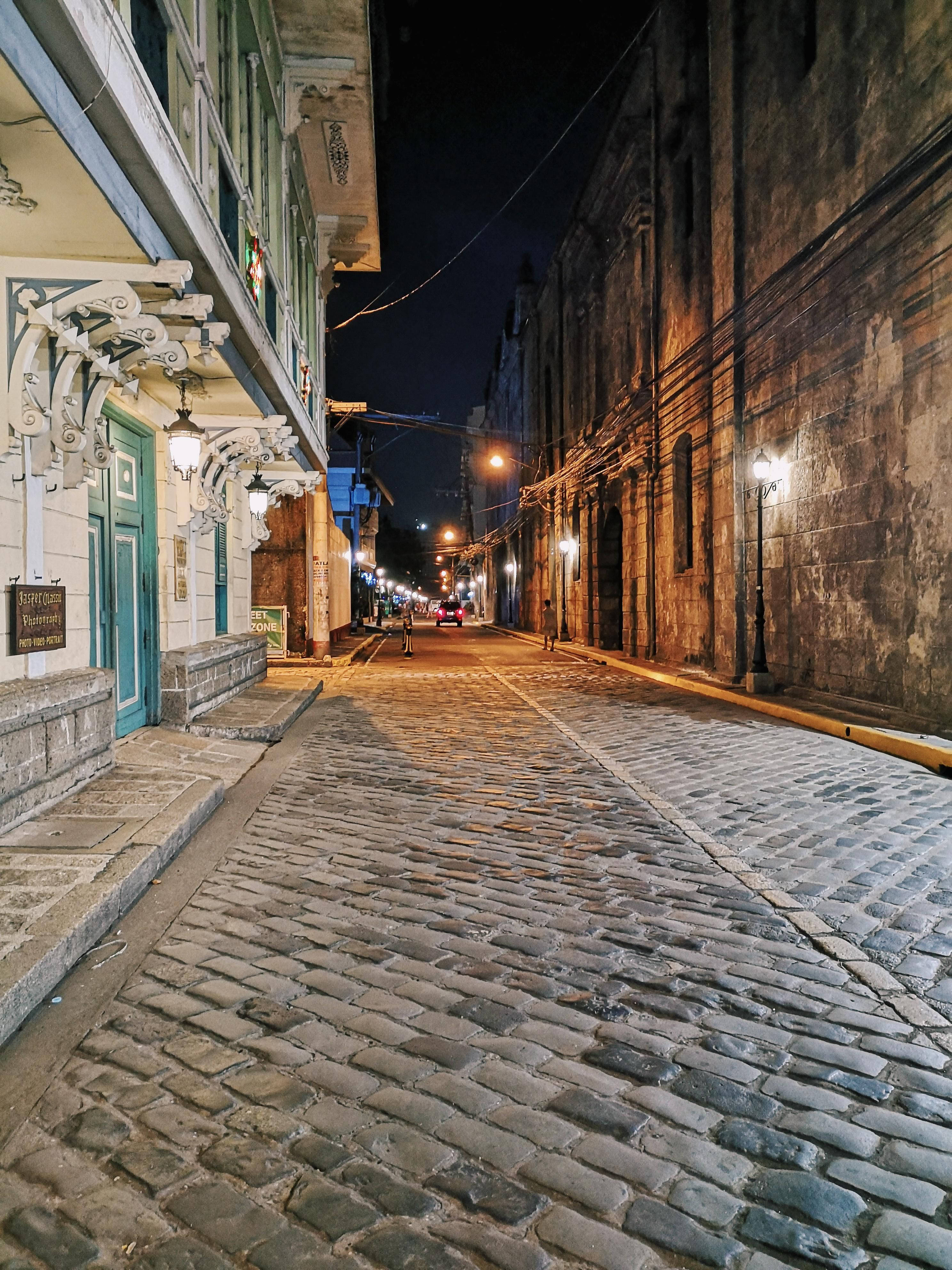 Manila 桌布 壁紙 — 一條狹窄的鵝卵石街道，夜晚被溫暖的路燈和復古建築外牆照亮。