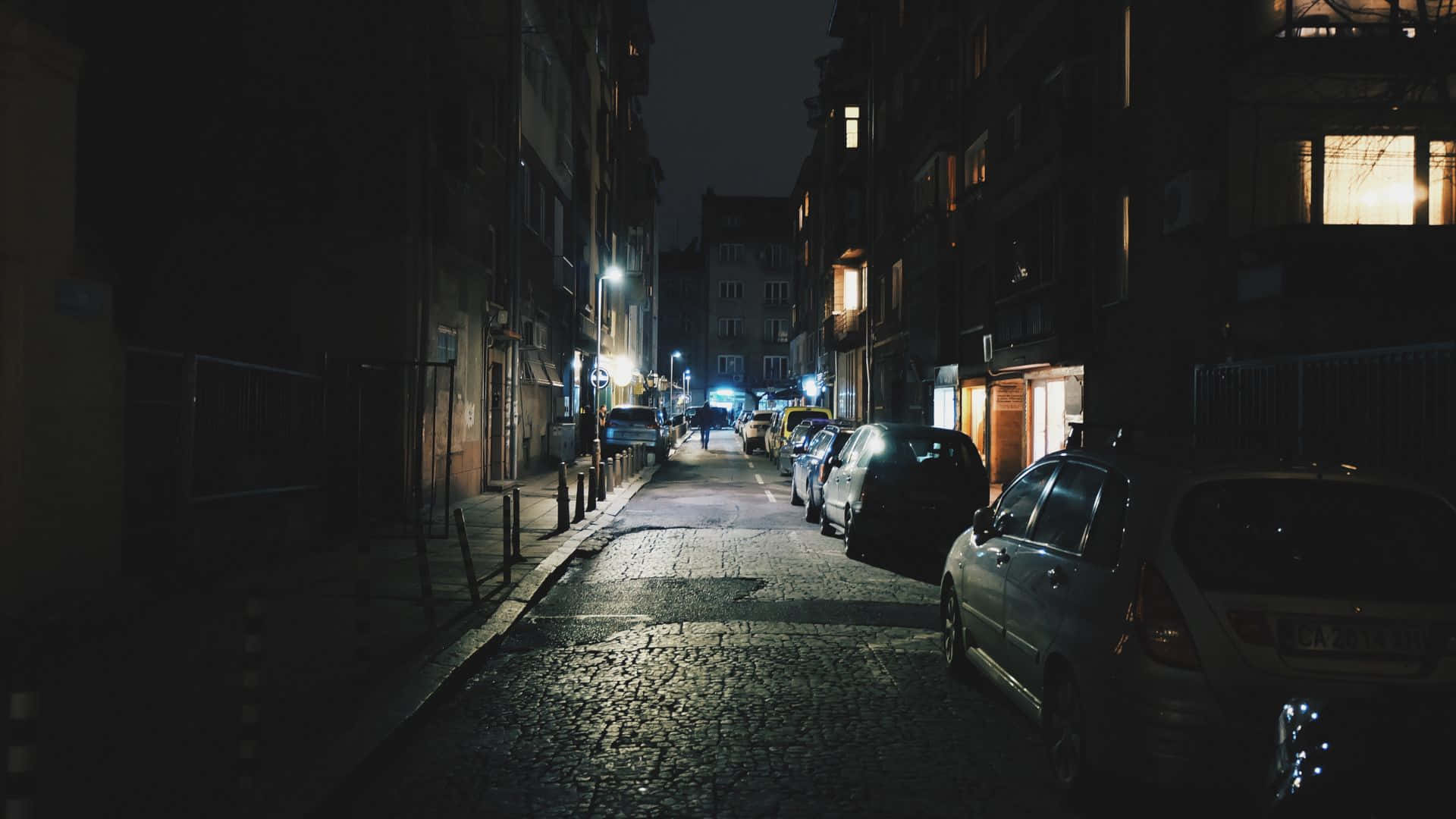 黑夜 桌布 壁紙 — 一條狹窄的鵝卵石街道在夜晚被街燈和溫暖的窗戶光芒照亮，兩旁停滿了汽車。
