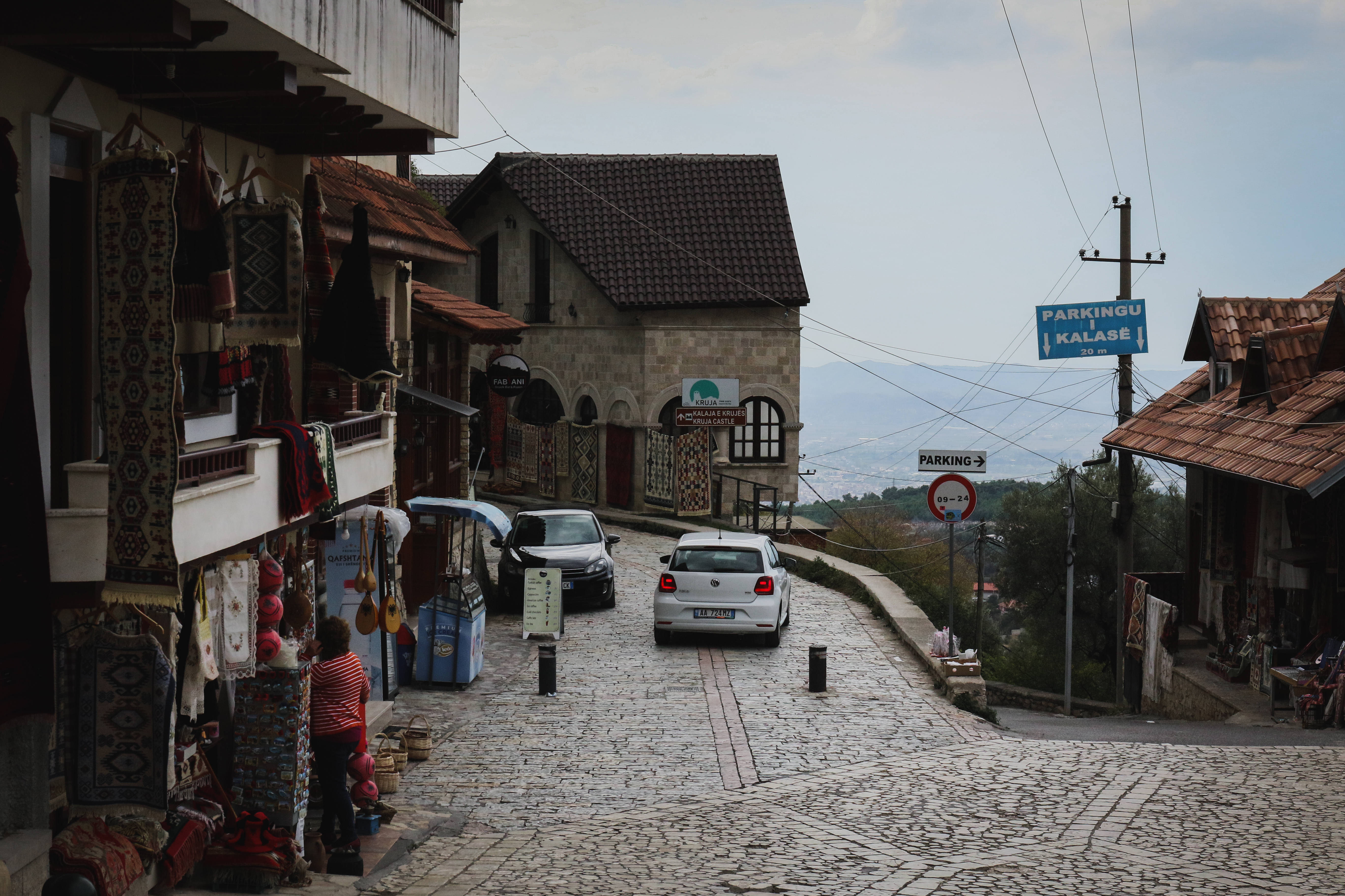 Albania 桌布 壁紙 — 一條狹窄的鵝卵石街道蜿蜒上山，穿過一個古色古香的村莊，村內有傳統的石頭建築和停泊的汽車，天空多雲。