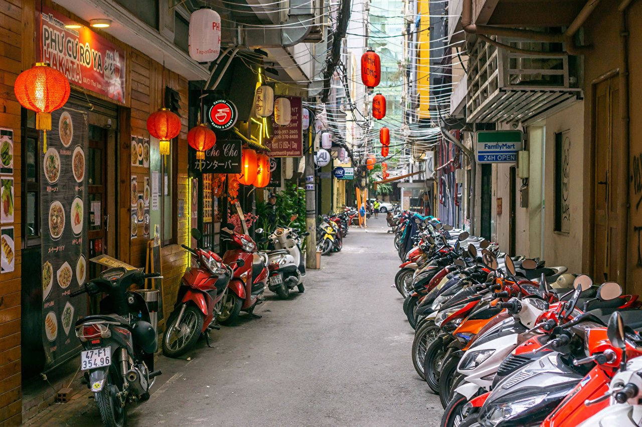 Hanoi 桌布 壁紙 — 一條狹窄而繁忙的小巷，兩旁停滿了機車，上方掛著發光的紅色燈籠，襯托出充滿活力的商店門面。