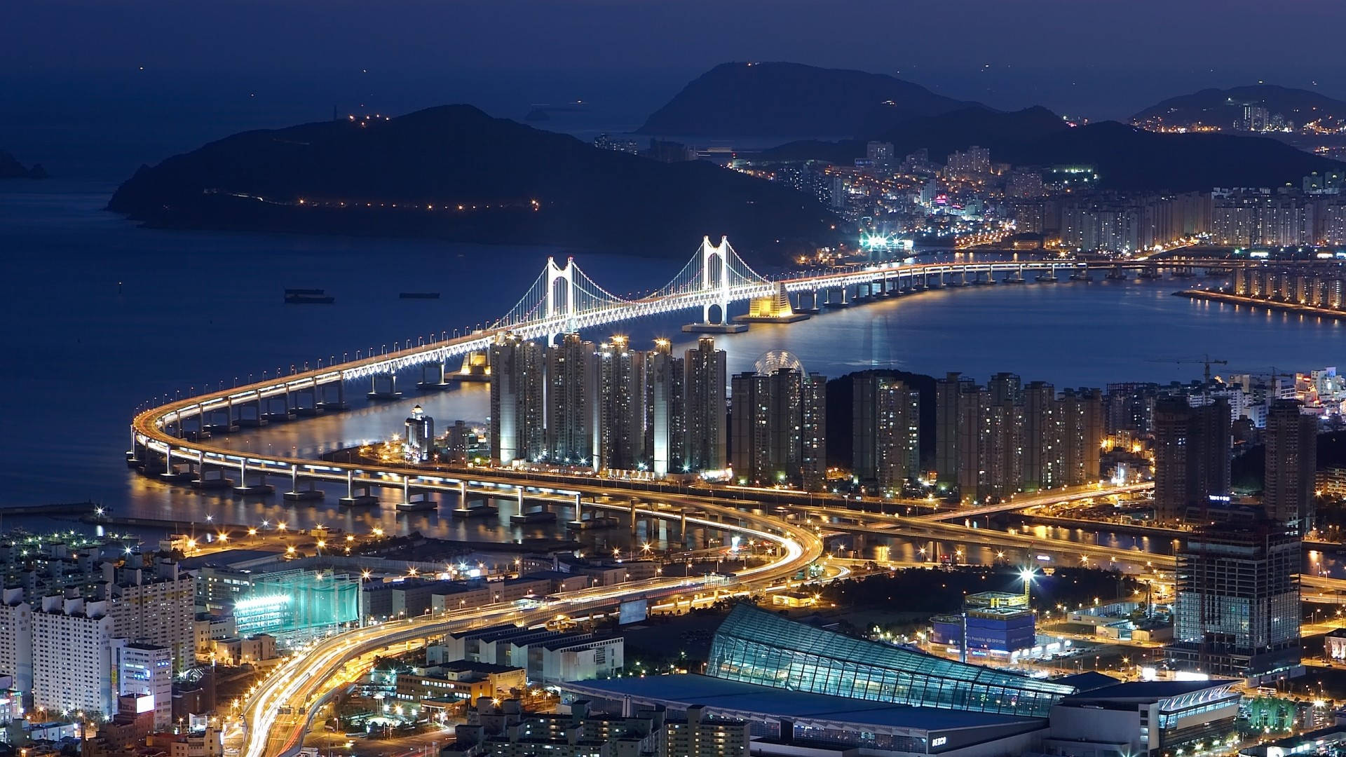 Busan 桌布 壁紙 — 一條發光的吊橋橫跨海灣，下方是燈火通明的城市景觀和蜿蜒的高速公路，壯麗的夜景。