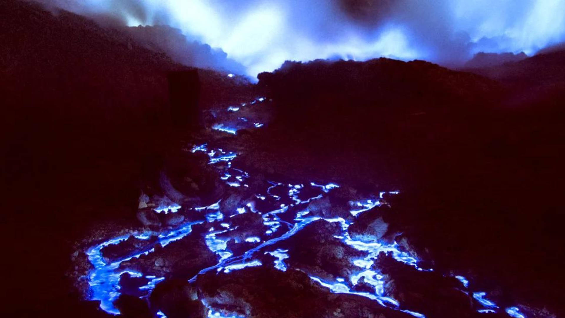 Flames 桌布 壁紙 — 一條發光的藍色熔岩河流經黑暗的火山景觀，天空風暴密佈。