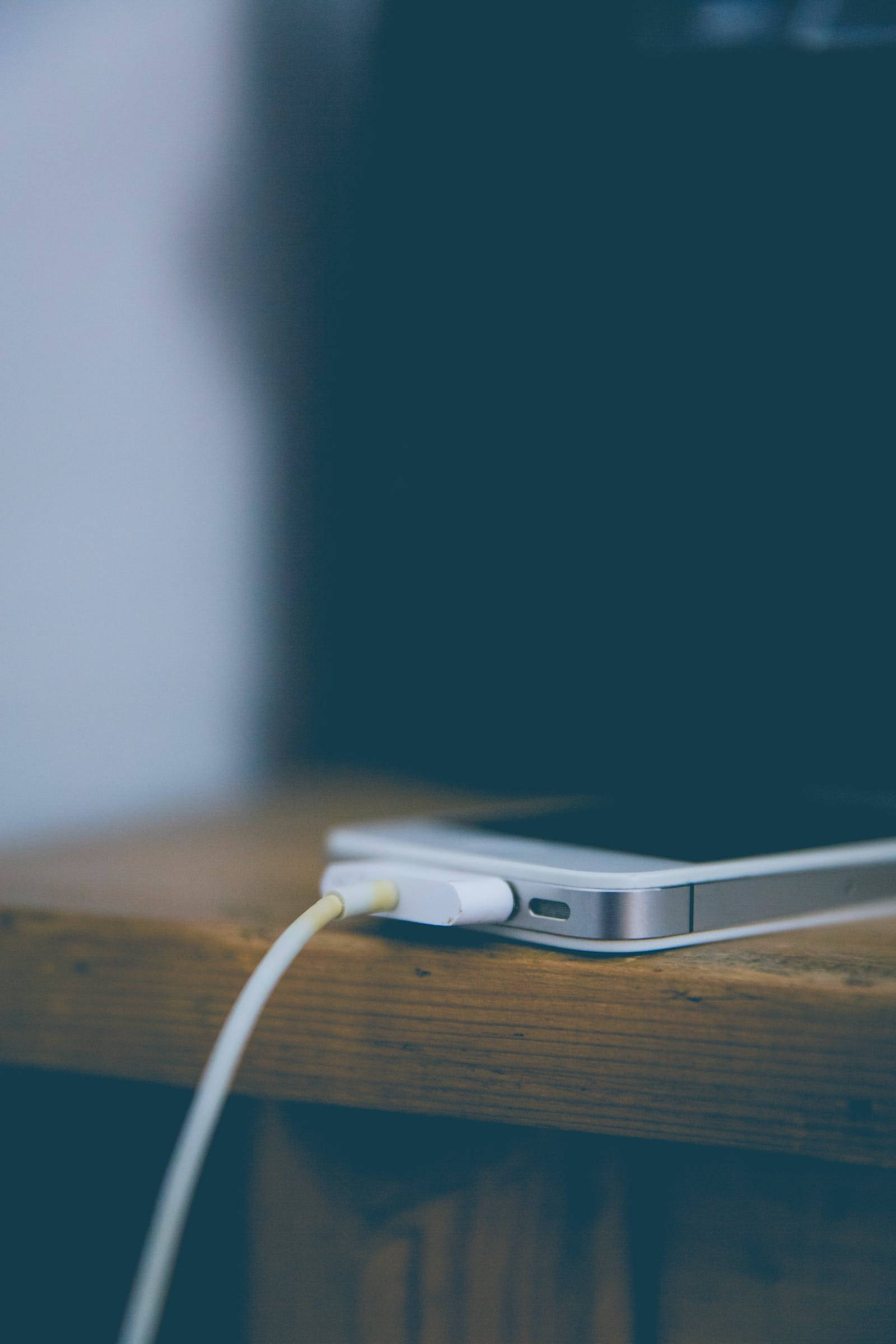 深色 iPhone 桌布 壁紙 — 一條白色手機充電線插入一部銀色智慧型手機，手機放在質樸的木質表面上。