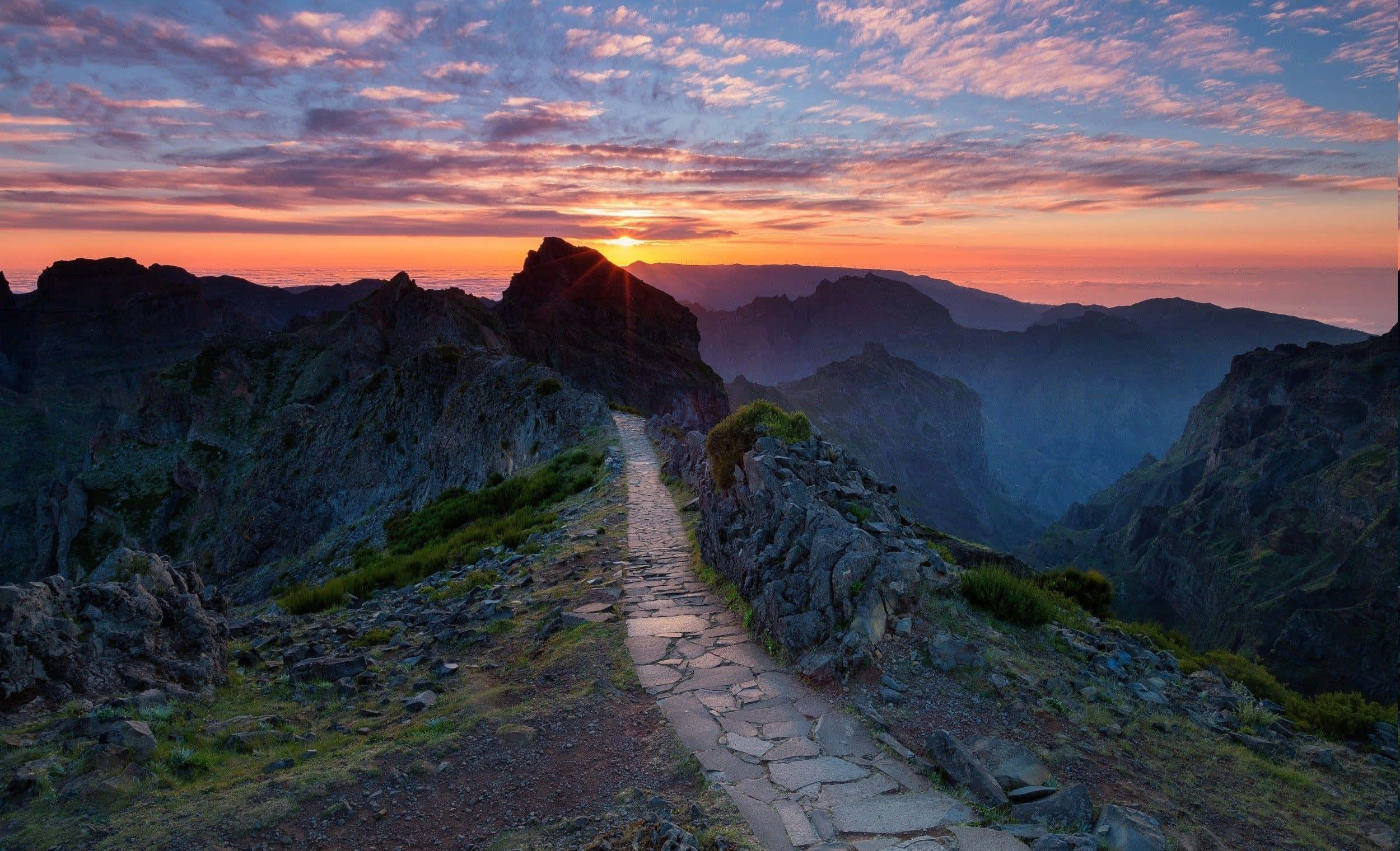 Hiking 桌布 壁紙 — 一條石徑蜿蜒穿過崎嶇的山脊，夕陽西下，上方是鮮豔的橙色和紫色雲彩。
