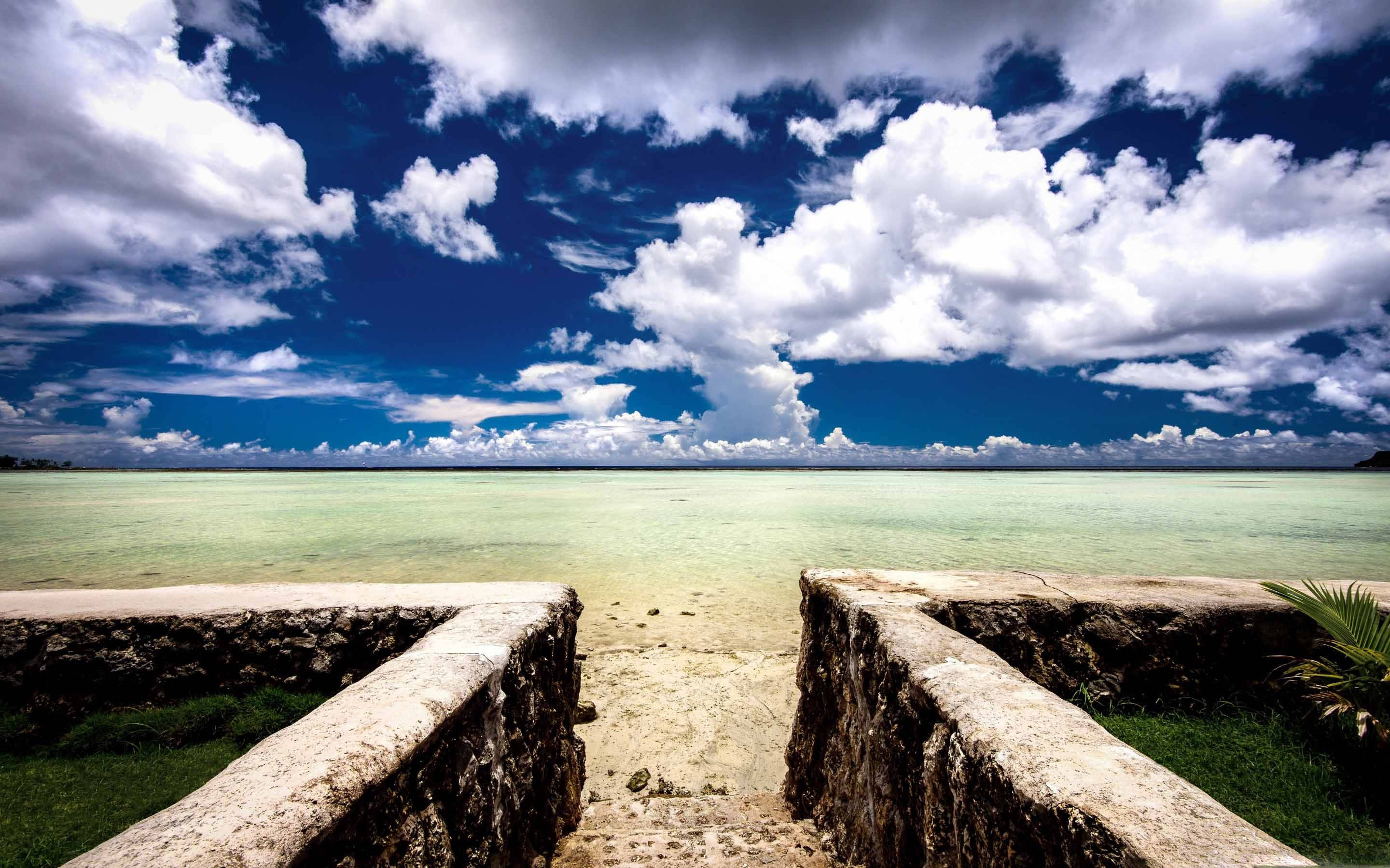 Guam 桌布 壁紙 — 一條石階通往寧靜的碧綠潟湖，上方是充滿蓬鬆白雲的鮮豔藍天。