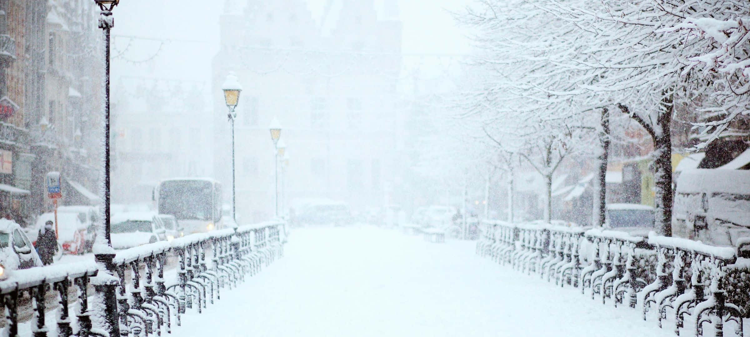 2400x1080 桌布 壁紙 — 一條積雪的城市街道，有被雪覆蓋的樹木和欄杆，在漫天大雪中，遠處的建築物模糊不清。