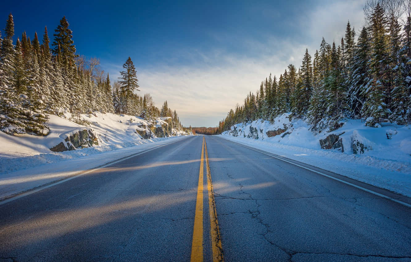 Road 桌布 壁紙 — 一條積雪的道路延伸至遠方，兩旁是常綠樹，在黃金時段清澈的藍天下。