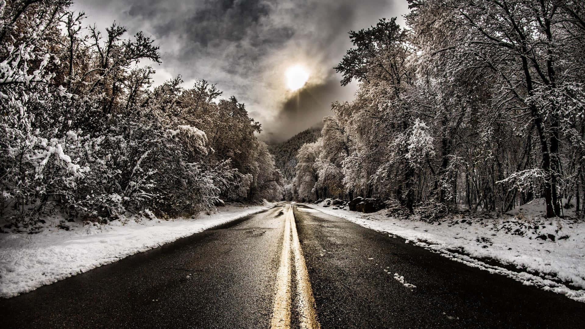Icy Road 桌布 壁紙 — 一條積雪的道路蜿蜒穿過霜凍的森林，天空多雲而戲劇化，明亮的陽光穿透雲層。