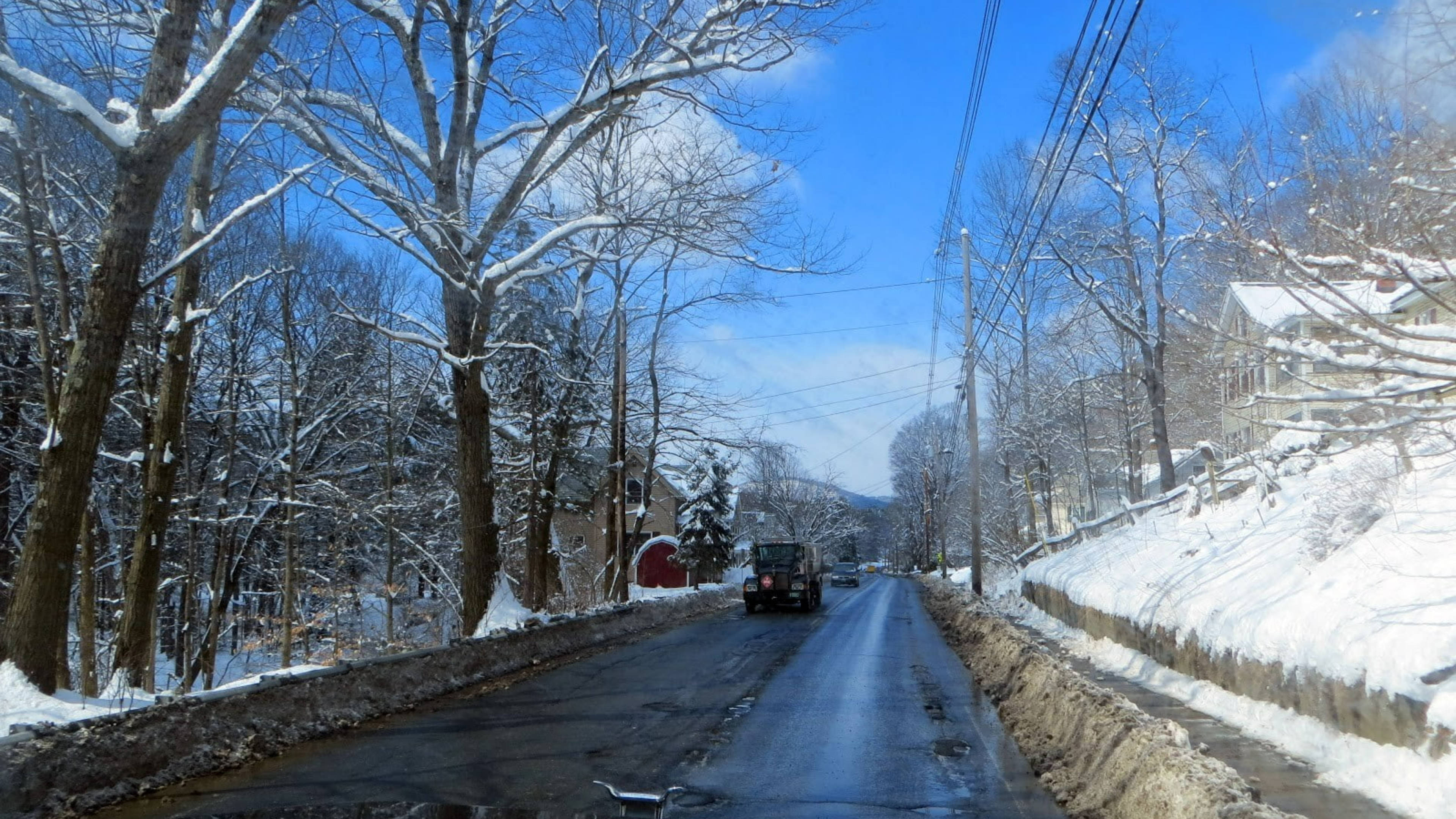 Vermont 桌布 壁紙 — 一條積雪鄉村道路蜿蜒穿過光禿的冬季樹木，在明亮的藍天下，一輛黑色車輛駛離。
