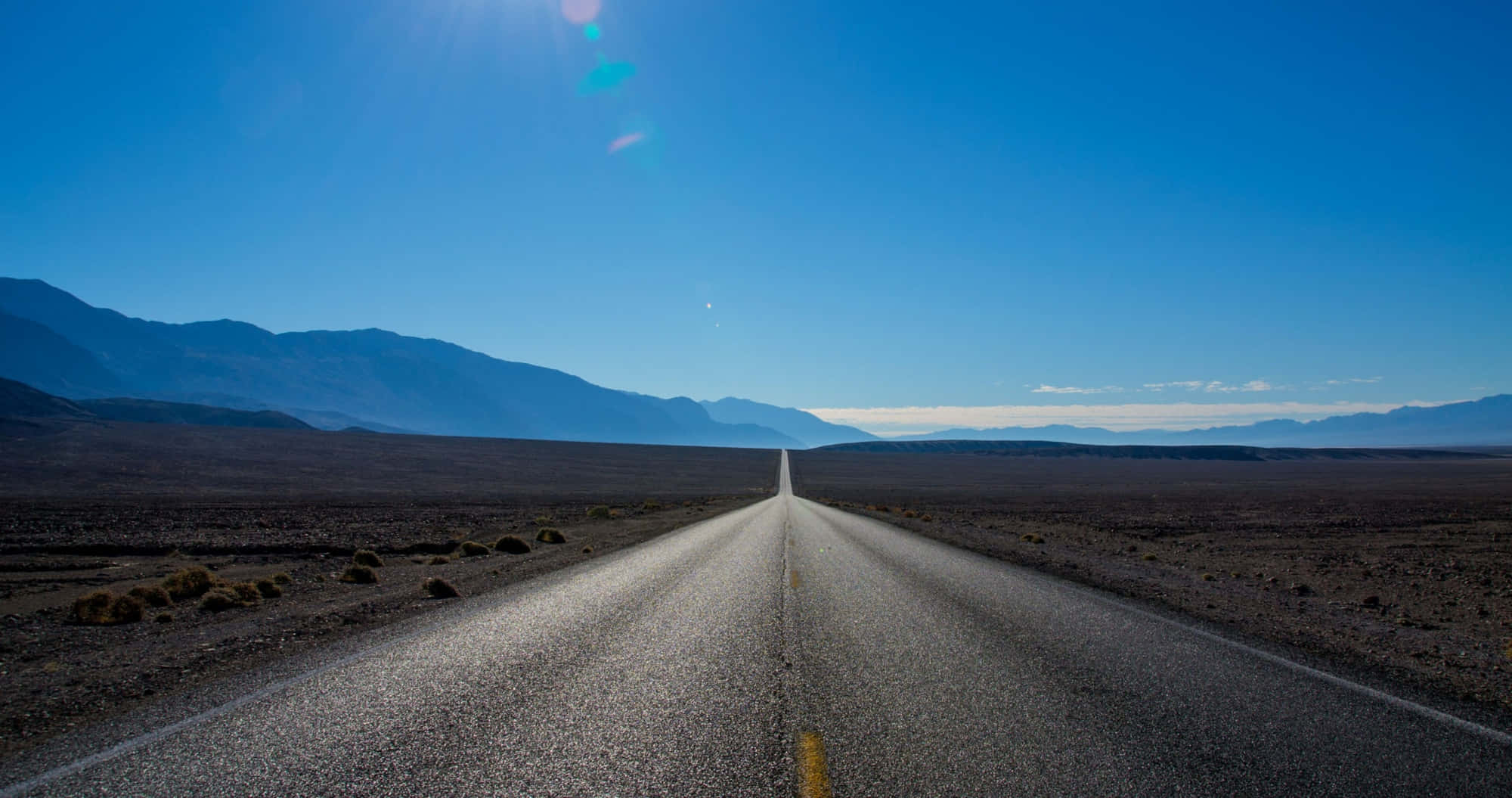 Highways 桌布 壁紙 — 一條筆直的柏油路在晴朗的藍天下，穿過荒蕪的沙漠景觀，一直延伸到遠山。