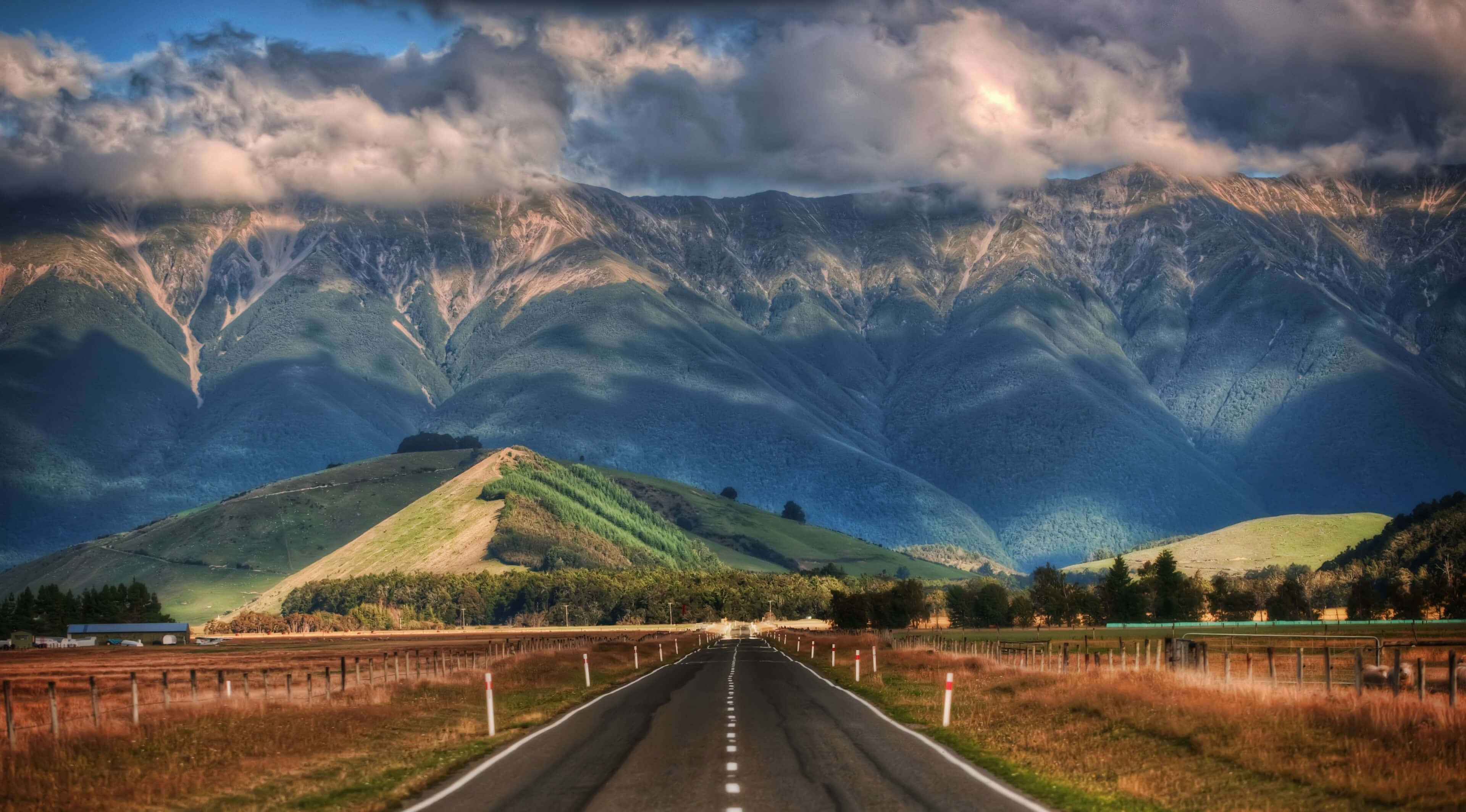 Road 桌布 壁紙 — 一條筆直的柏油路延伸向高聳的山脈，天空多雲而戲劇性，陽光穿透雲層。