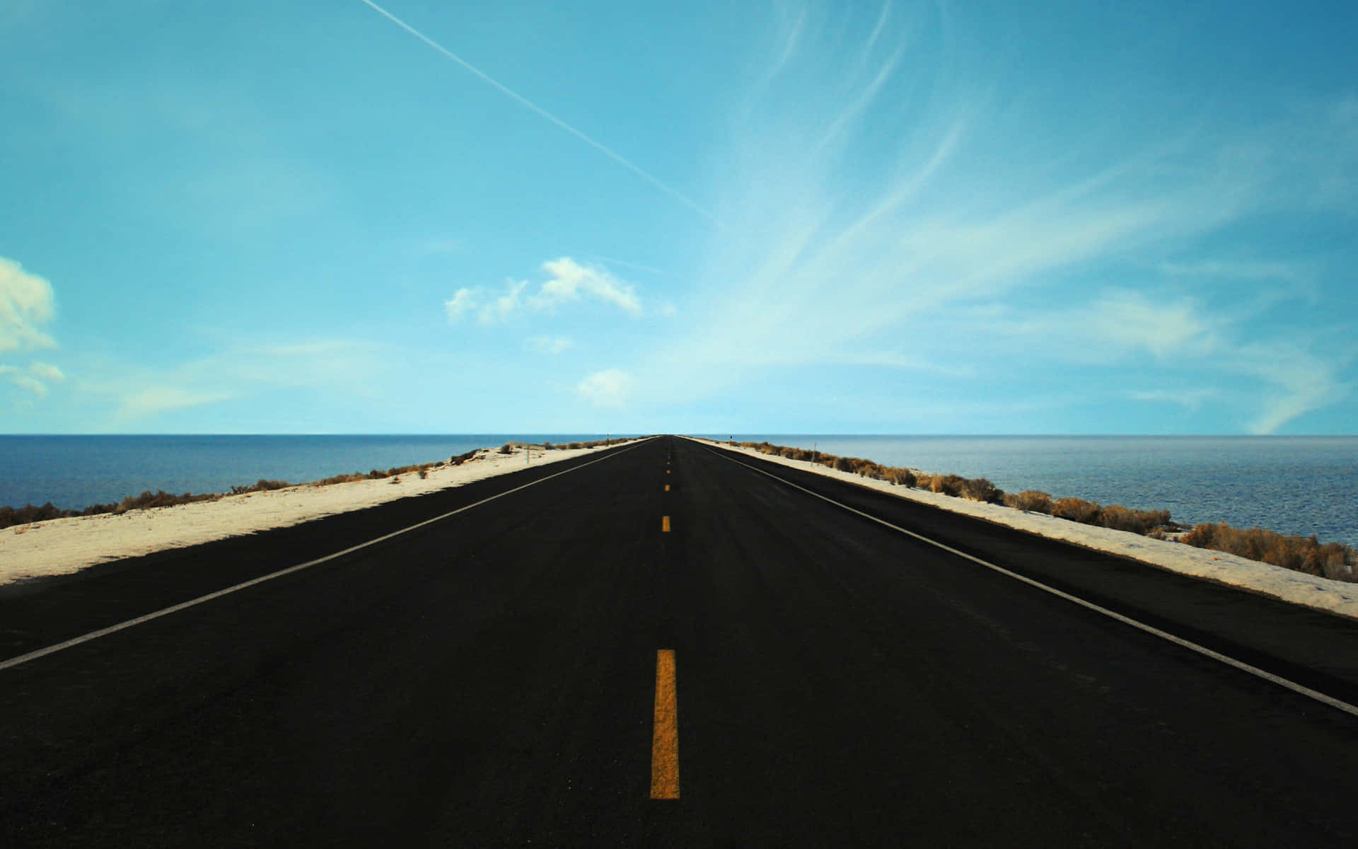 Runway 桌布 壁紙 — 一條筆直的柏油路延伸至地平線，兩旁是海洋，上方是廣闊的藍天，點綴著縷縷薄雲。