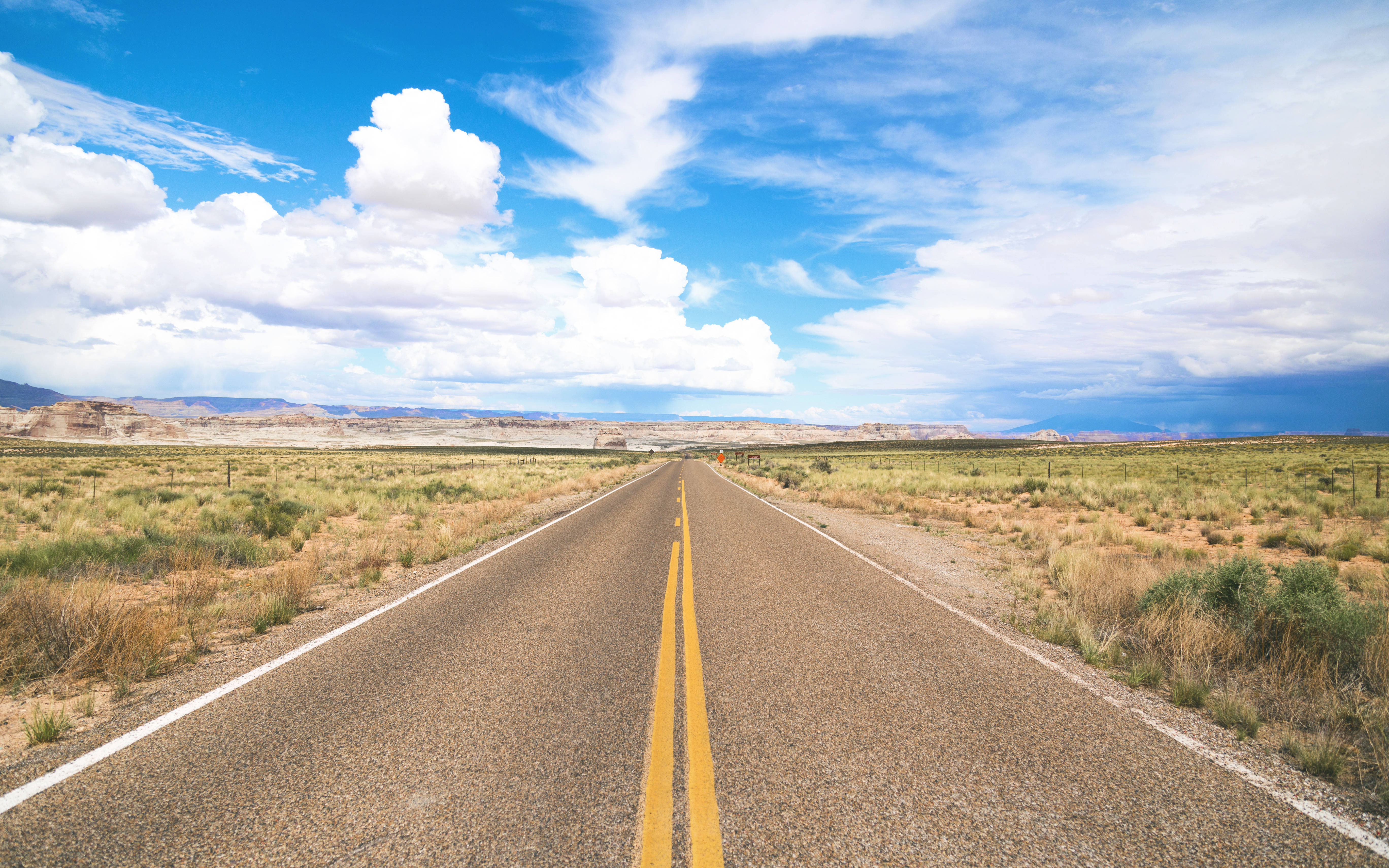 Road 桌布 壁紙 — 一條筆直的柏油路穿過廣闊乾旱的景觀，在明亮的藍天和蓬鬆的白雲下延伸至遠方。