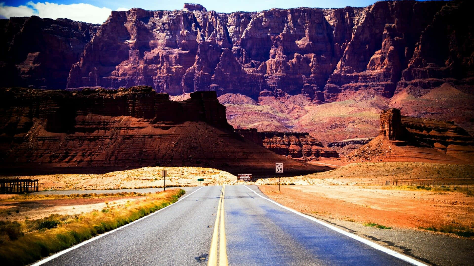 Arizona 桌布 壁紙 — 一條筆直的柏油道路在明亮的藍天下延伸向高聳的紅色岩壁。
