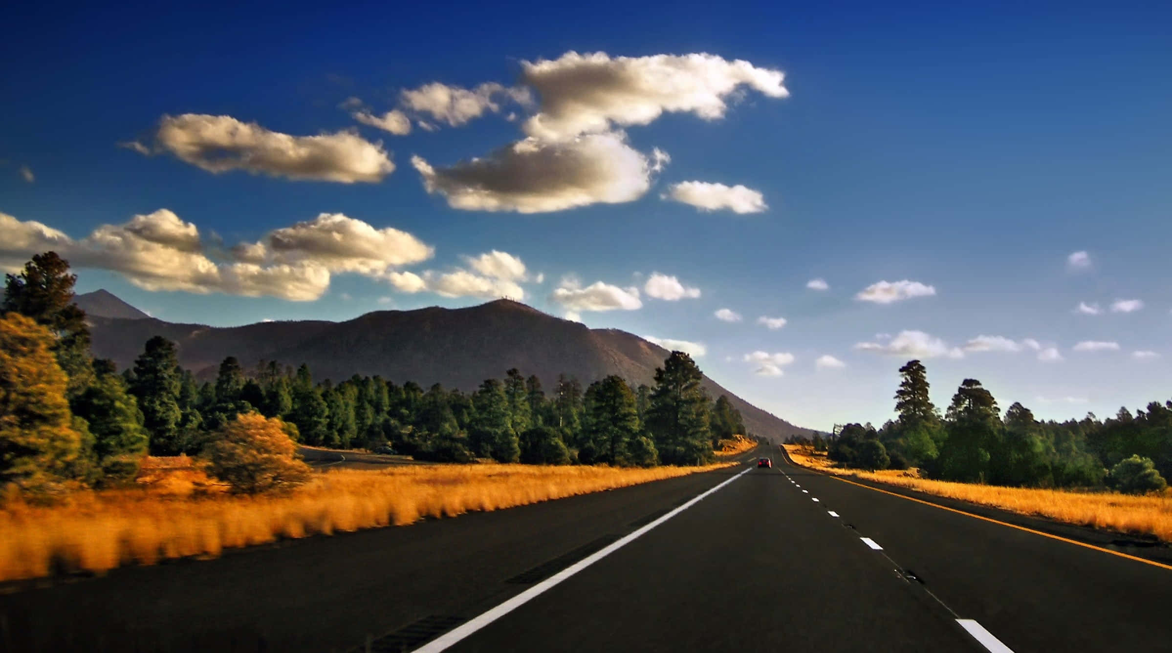 Highways 桌布 壁紙 — 一條筆直的柏油道路在明亮的藍天和散落的蓬鬆雲朵下，延伸向遠方的山脈。