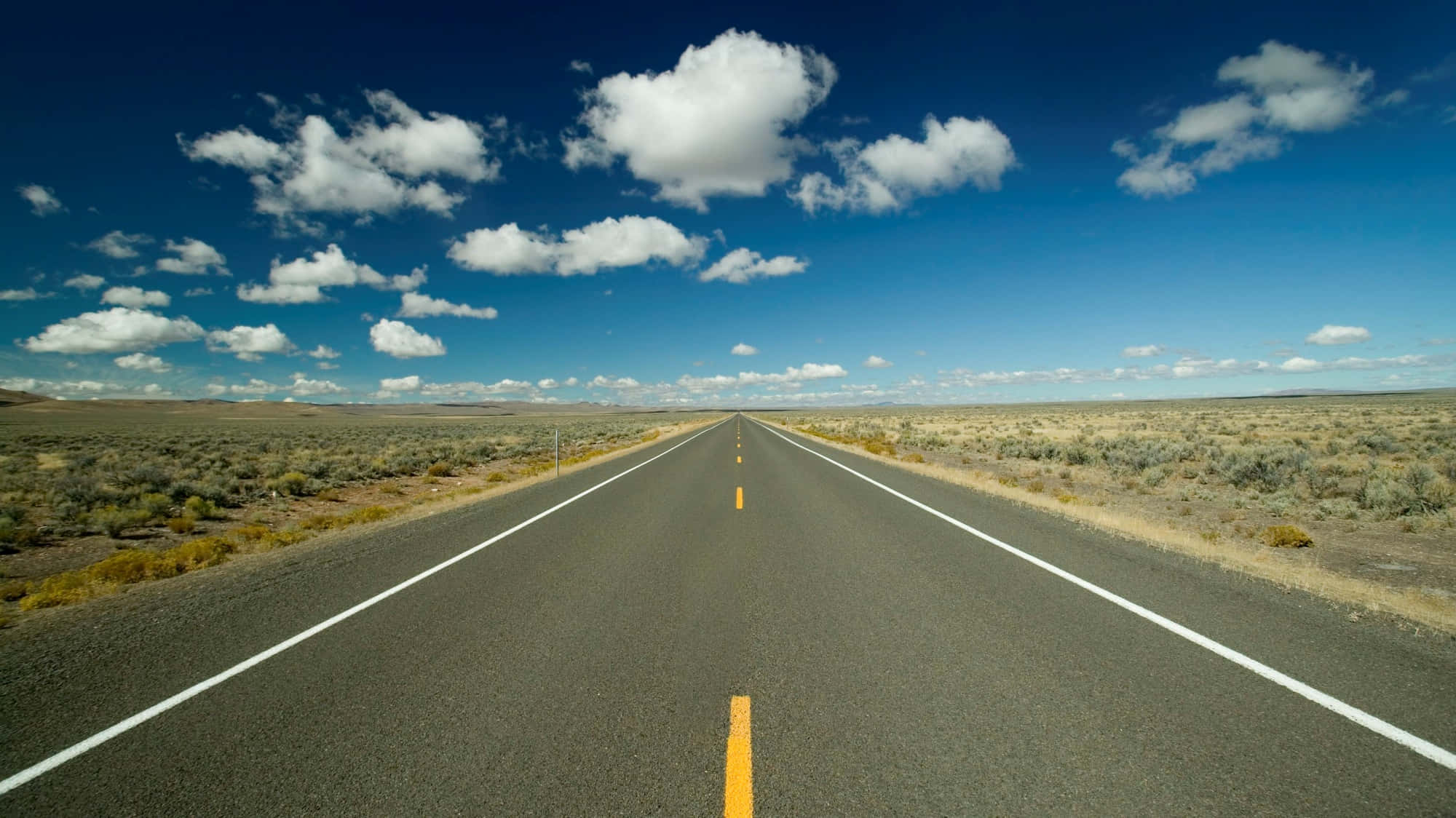 Highways 桌布 壁紙 — 一條筆直的柏油道路在晴朗藍天和蓬鬆白雲點綴下，無盡地延伸穿過乾燥的沙漠。