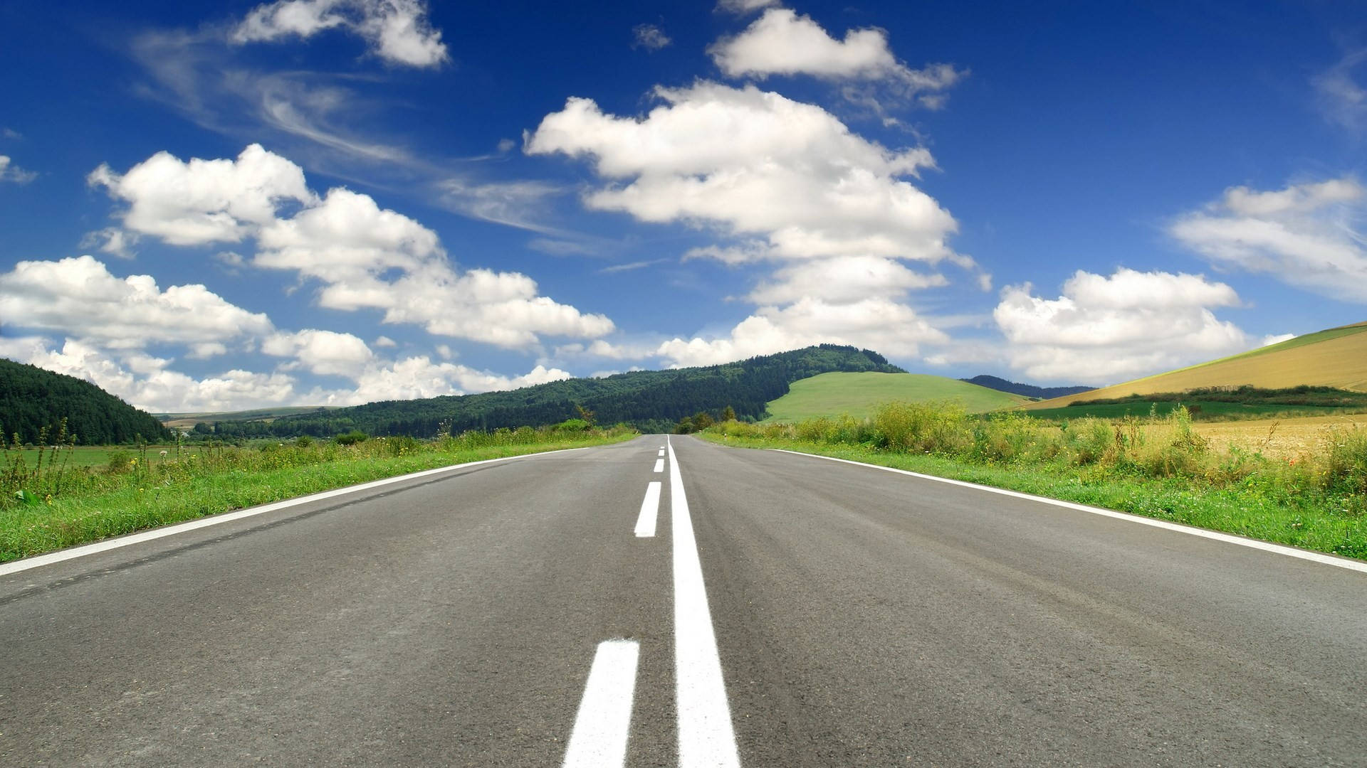 Country Road 桌布 壁紙 — 一條筆直的柏油道路在晴朗藍天和蓬鬆白雲下，伸向遠方的綠色山丘。