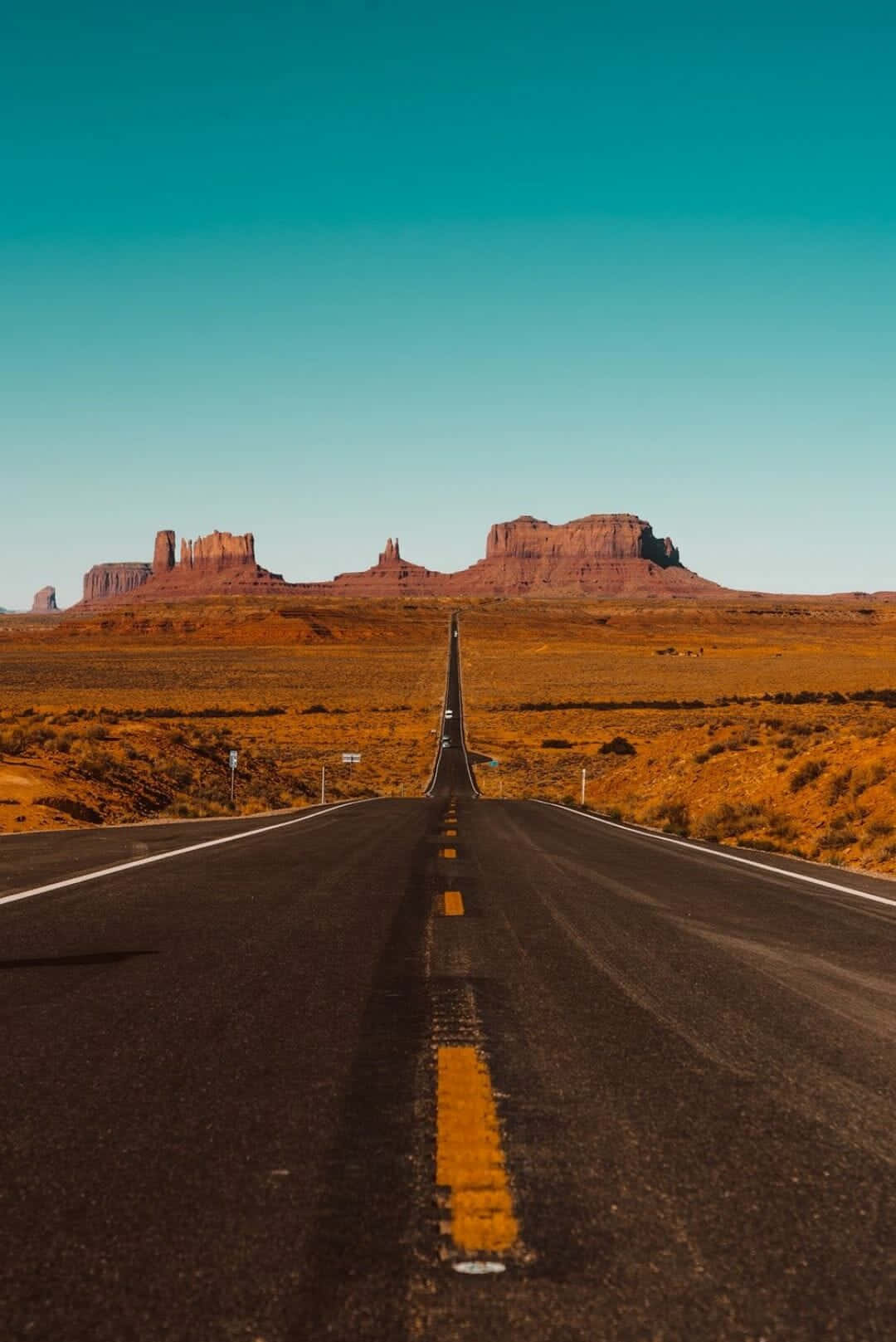 Desert iPhone 桌布 壁紙 — 一條筆直的沙漠高速公路在清澈的藍綠色天空下延伸向標誌性的紅色岩丘。