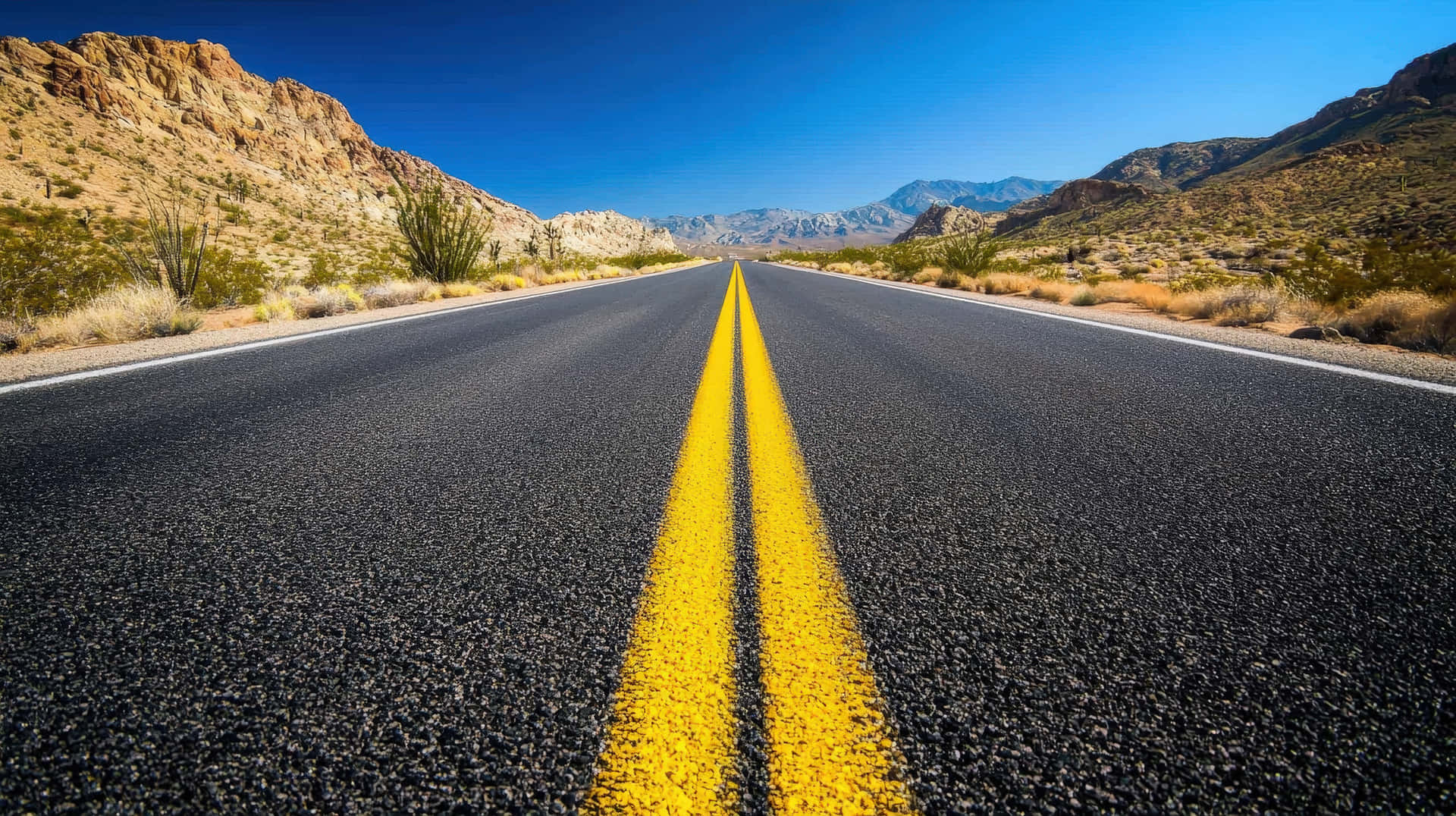 Long desert highway with yellow dividing lines stretching towards distant mountains 壁紙 — 一條筆直的瀝青公路，帶有粗黃色線條，在晴朗藍天下延伸向遠山，處於沙漠景觀中。