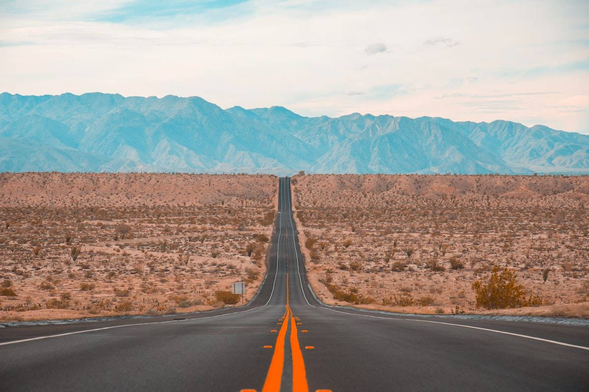 Road 桌布 壁紙 — 一條筆直的瀝青公路帶有橙色線條，在柔和的多雲天空下延伸至遠方的藍色山脈。