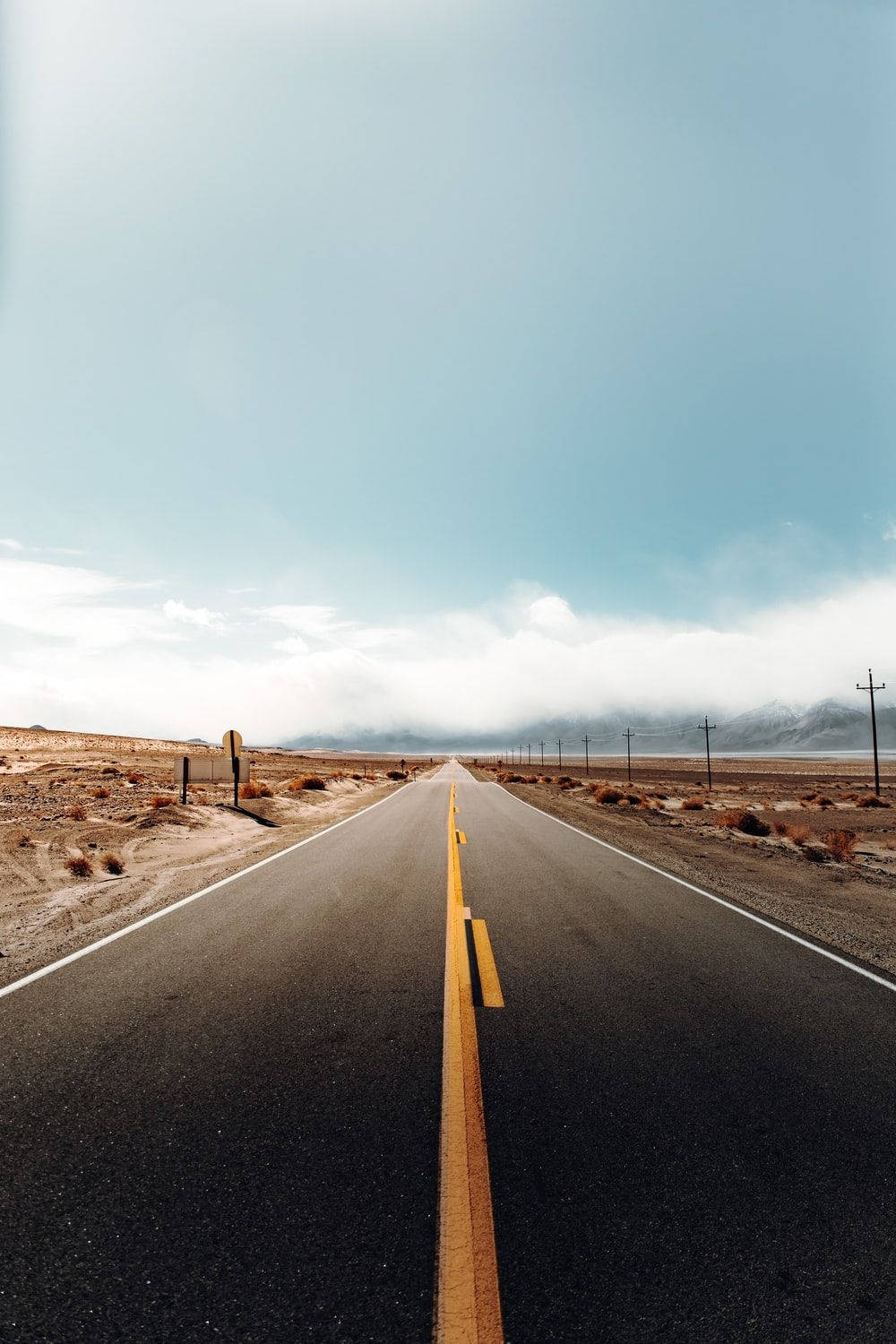 road trip 桌布 壁紙 — 一條筆直的瀝青公路穿過荒蕪的沙漠景觀，在廣闊的藍天下延伸至遠方，遠處有山脈。
