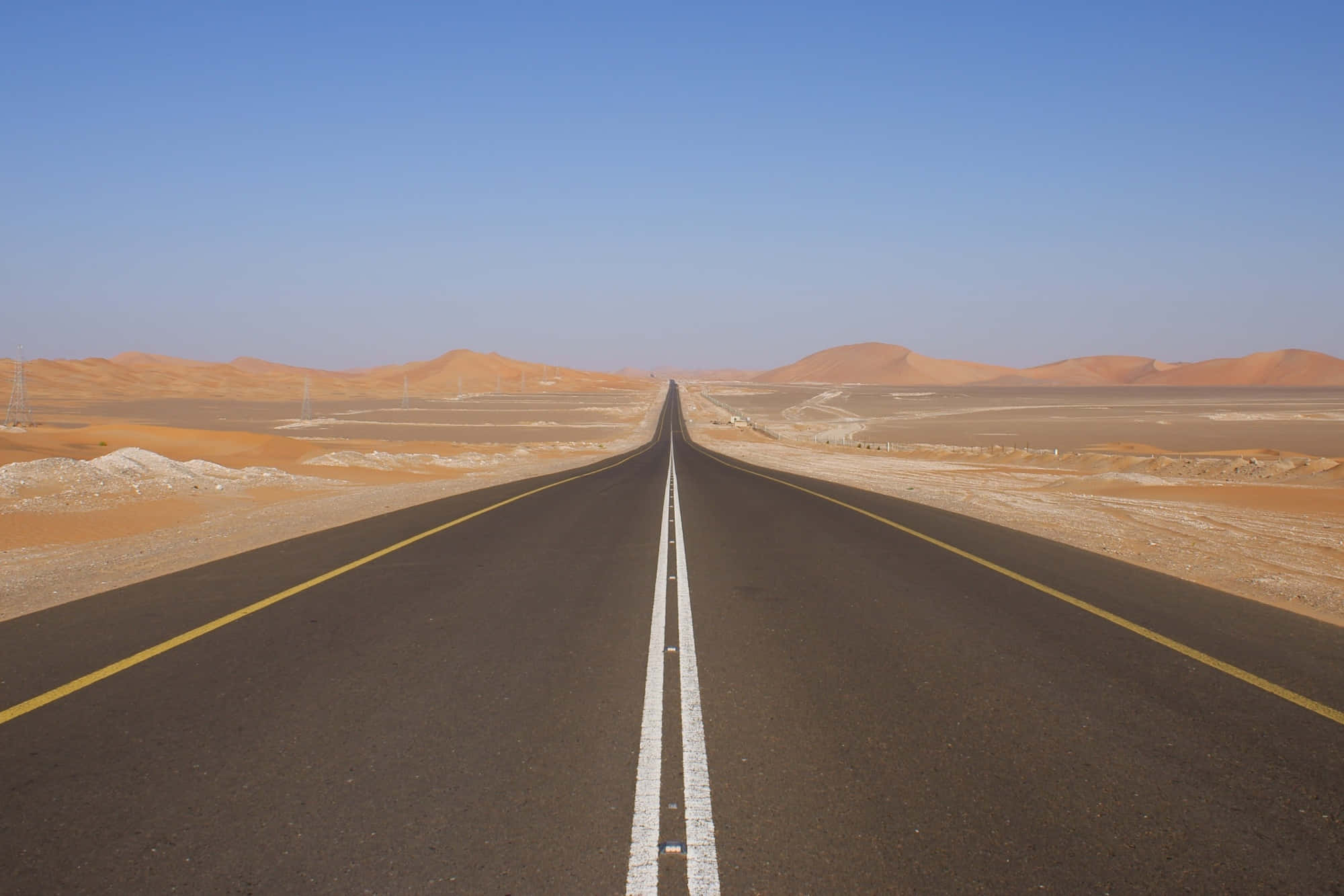 Highways 桌布 壁紙 — 一條筆直的瀝青路在晴朗的藍天下無盡地延伸穿過廣闊的沙漠，兩側遠處有沙丘。