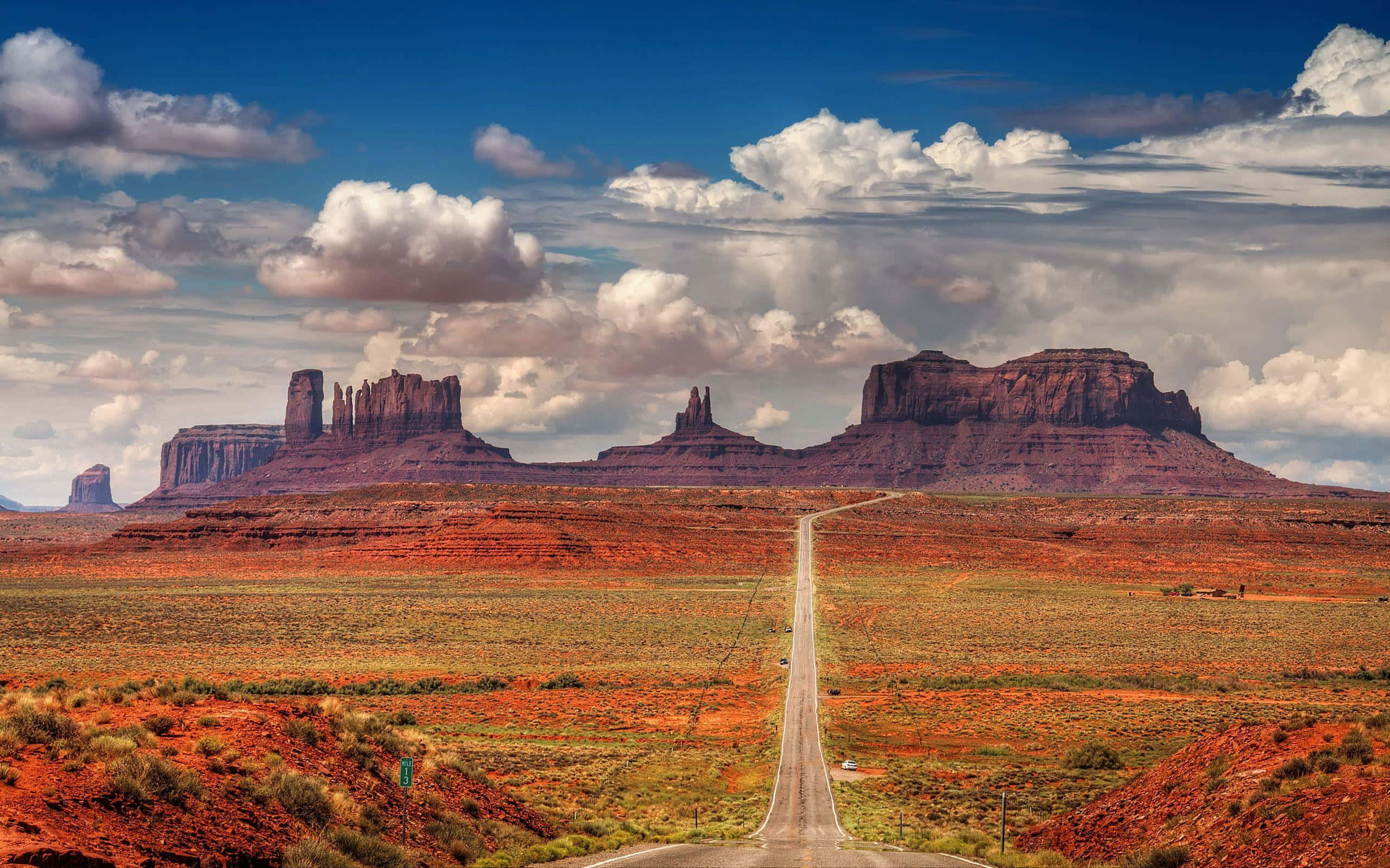 Arizona 桌布 壁紙 — 一條筆直的道路在廣闊的沙漠景觀中，延伸向高聳的紅色岩石台地，上方是戲劇性的多雲天空。