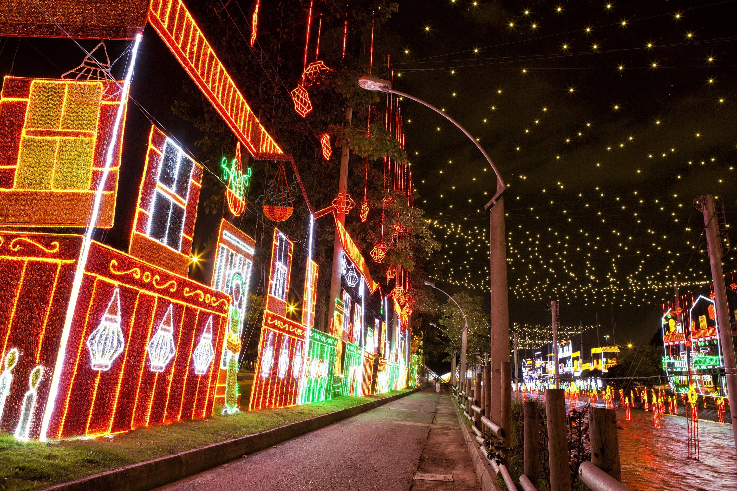 Medellin 桌布 壁紙 — 一條節日夜晚的街道，點綴著鮮豔的紅、綠、黃燈光，照亮建築物和懸掛的燈籠，在星空下。