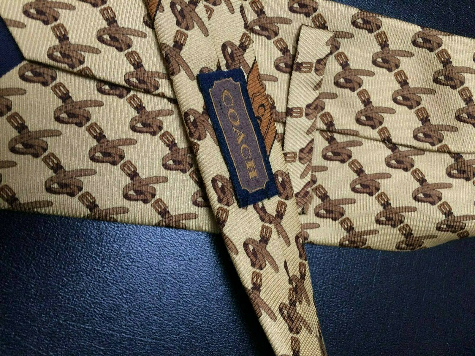 Coach 桌布 壁紙 — 一條米色Coach領帶帶有棕色交錯G圖案，折疊放在有紋理的藍色皮革表面上。