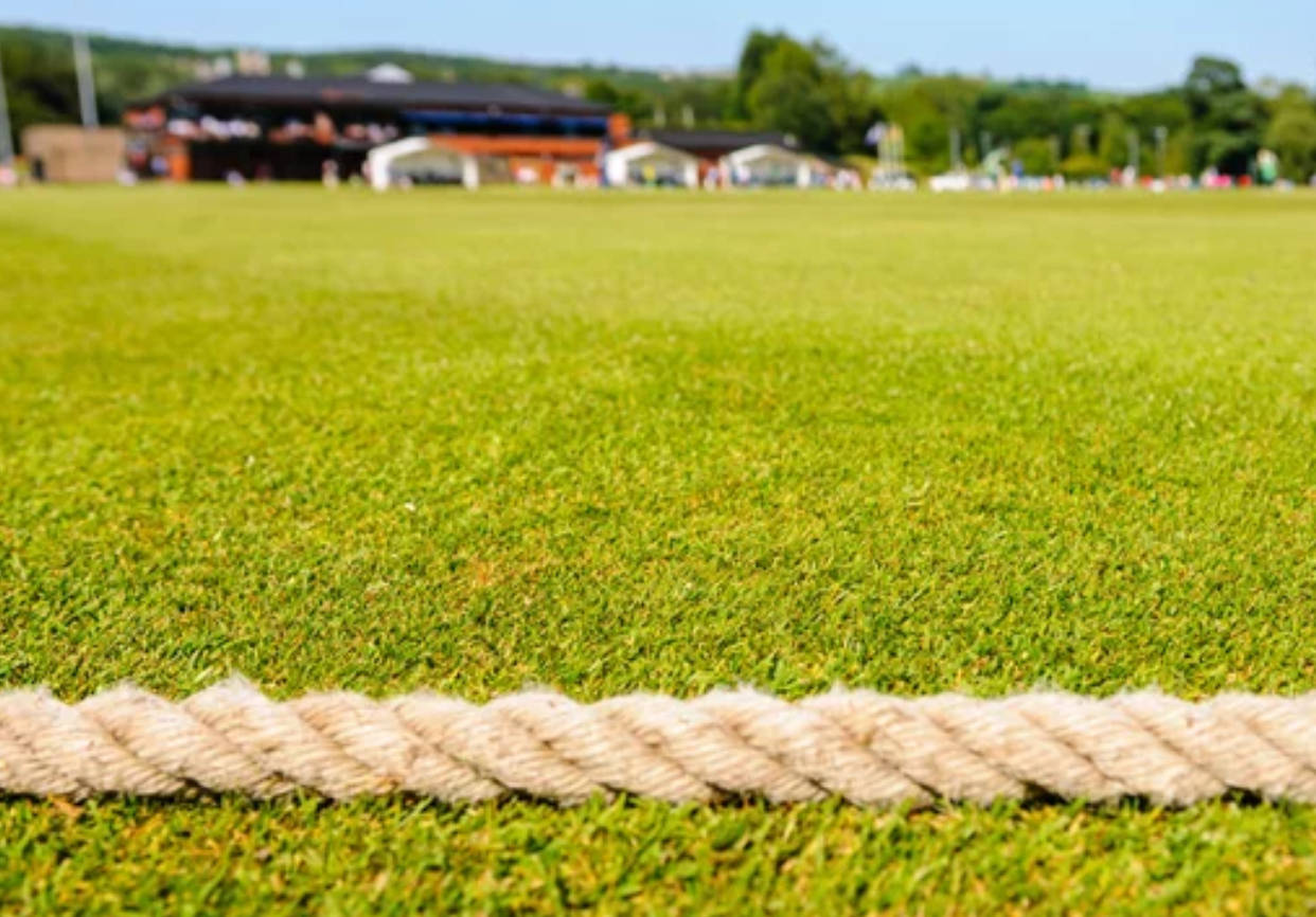 Cricket Ground 桌布 壁紙 — 一條粗米色繩索橫跨在鮮綠色草地上，背景是模糊的運動場和看台。