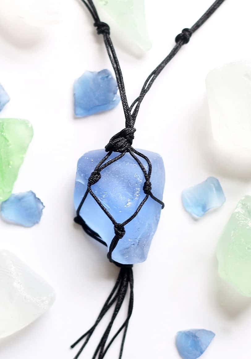 Sea Glass 桌布 壁紙 — 一條精緻的黑色繩結項鍊，配以藍色海玻璃吊墜，放置在明亮的白色表面上，周圍散落著海玻璃碎片。