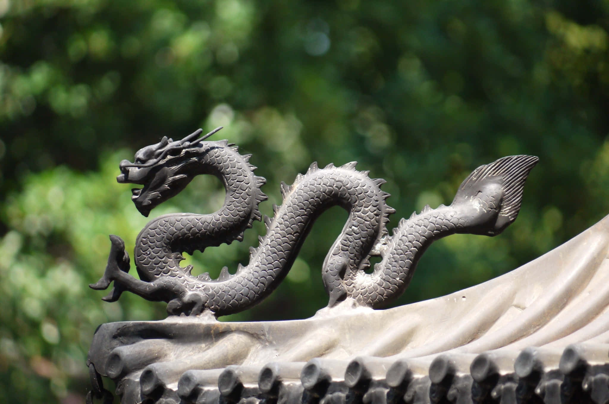 龍 桌布 壁紙 — 一條細節豐富的灰色龍雕塑優雅地盤旋在傳統瓦片屋頂上，背景是柔和的綠色樹葉。