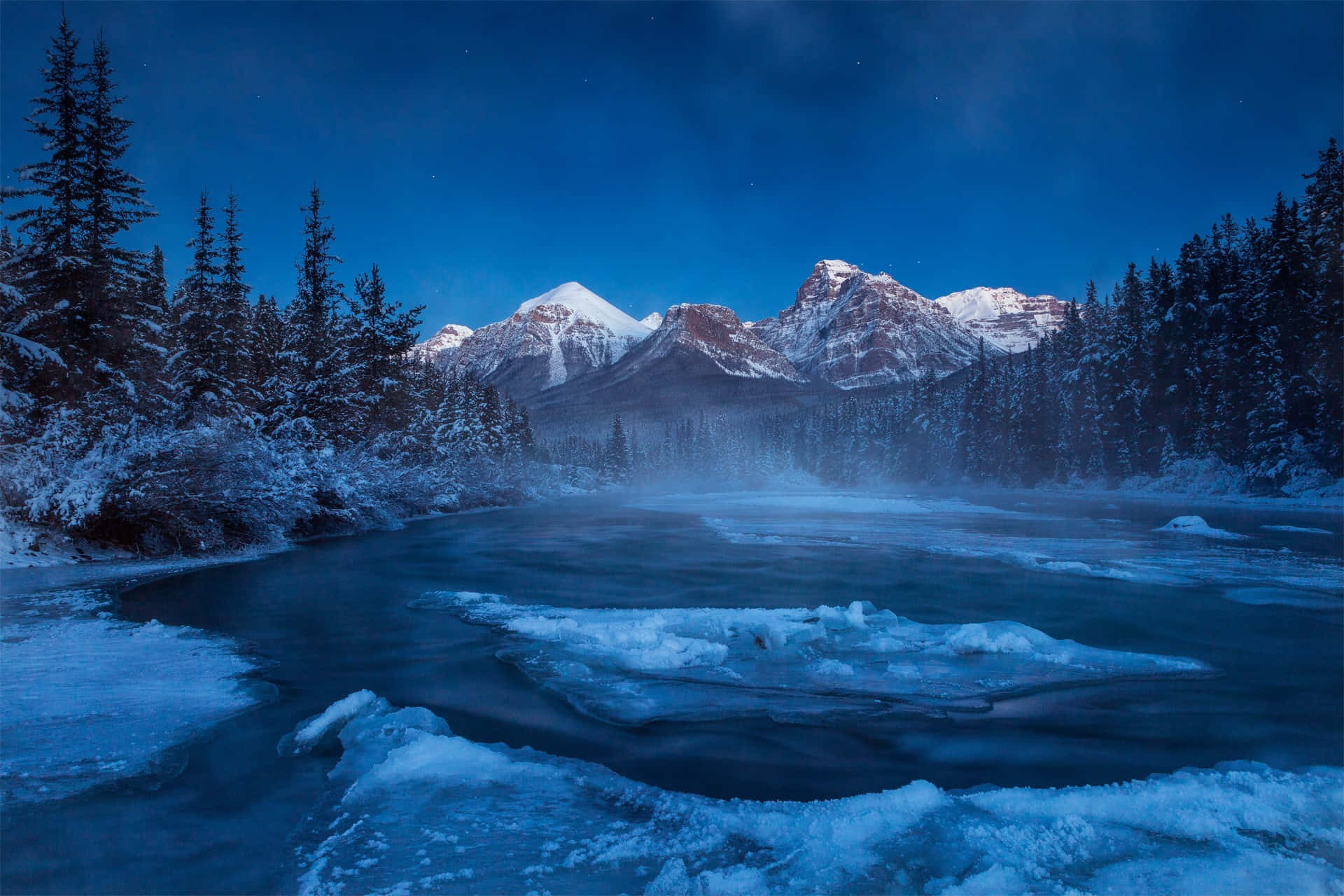  壁紙 — 一條結冰的河流蜿蜒穿過白雪覆蓋的松樹林，在繁星點點的冬夜天空下，遠處是積雪的山峰。