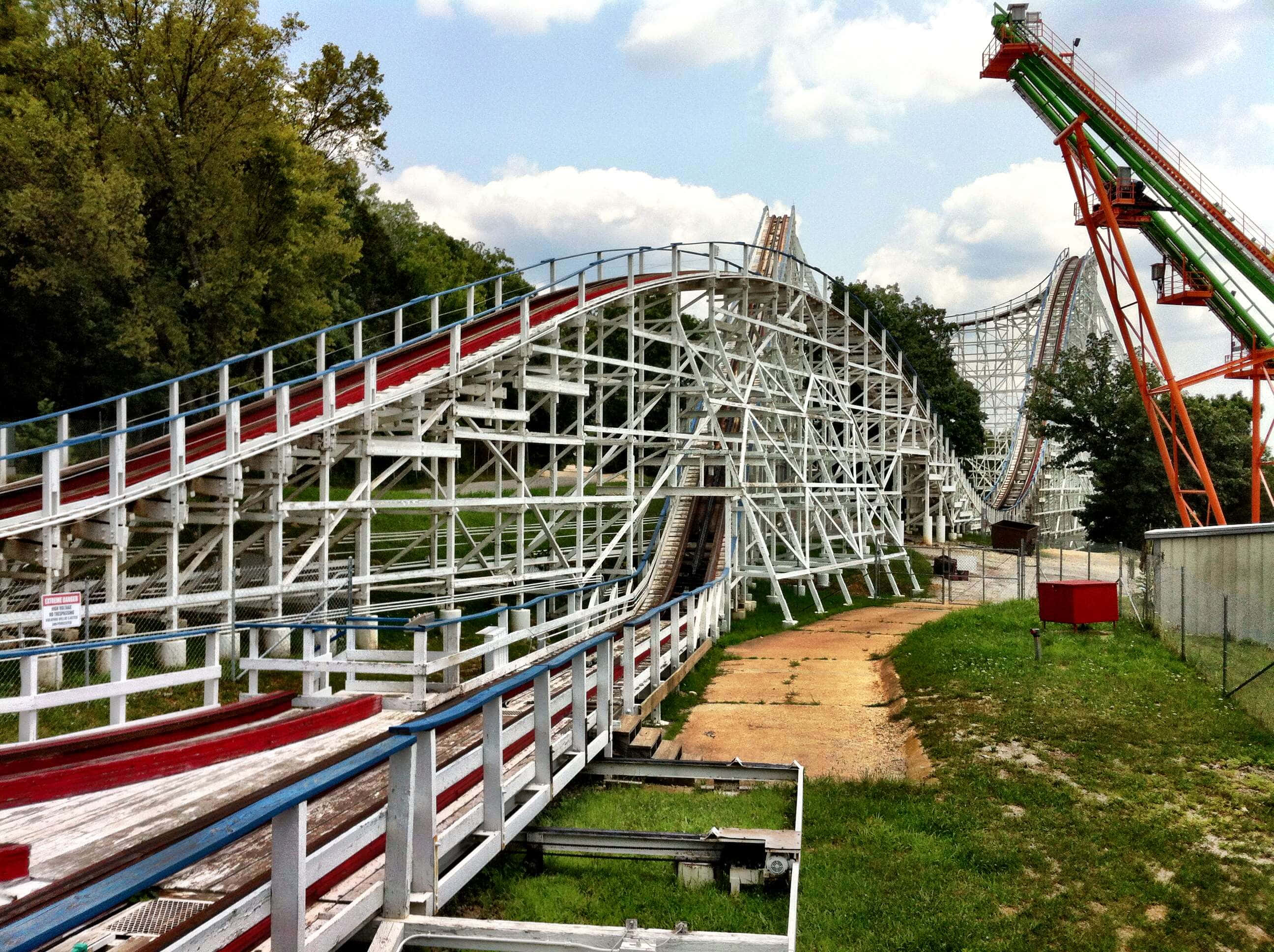 Roller Coaster 桌布 壁紙 — 一條經過風化的木製雲霄飛車，帶有紅色軌道和白色支架，在部分多雲的藍天下蜿蜒穿過一個草地公園。