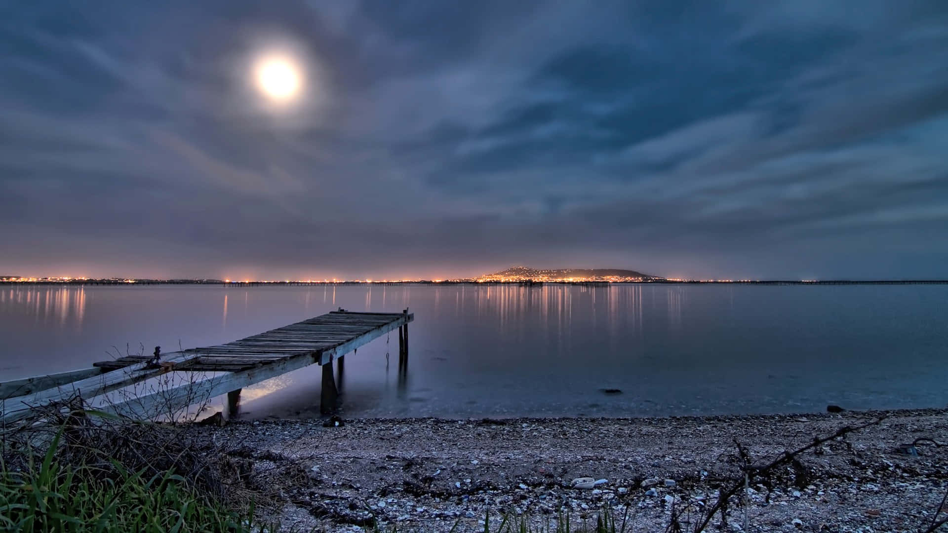 Beach At Night 桌布 壁紙 — 一條經風吹雨打的木碼頭延伸到平靜的湖面上，明亮的滿月下，城市燈光倒映在水中。