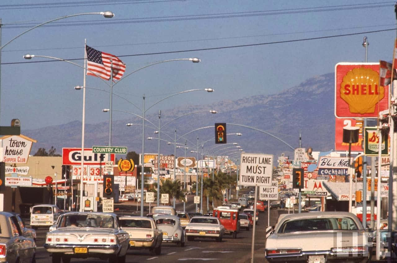 1970s 桌布 壁紙 — 一條繁忙的1960年代美國街道，充滿復古汽車和色彩繽紛的加油站招牌，在晴朗藍天和遠處山脈之下。