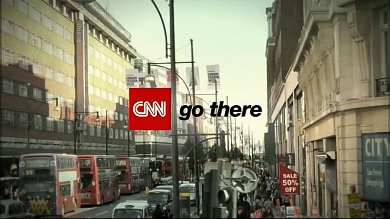 Cnn 桌布 壁紙 — 一條繁忙的城市街道，充滿紅色雙層巴士和行人，天空朦朧，帶有CNN go there標誌覆蓋層。